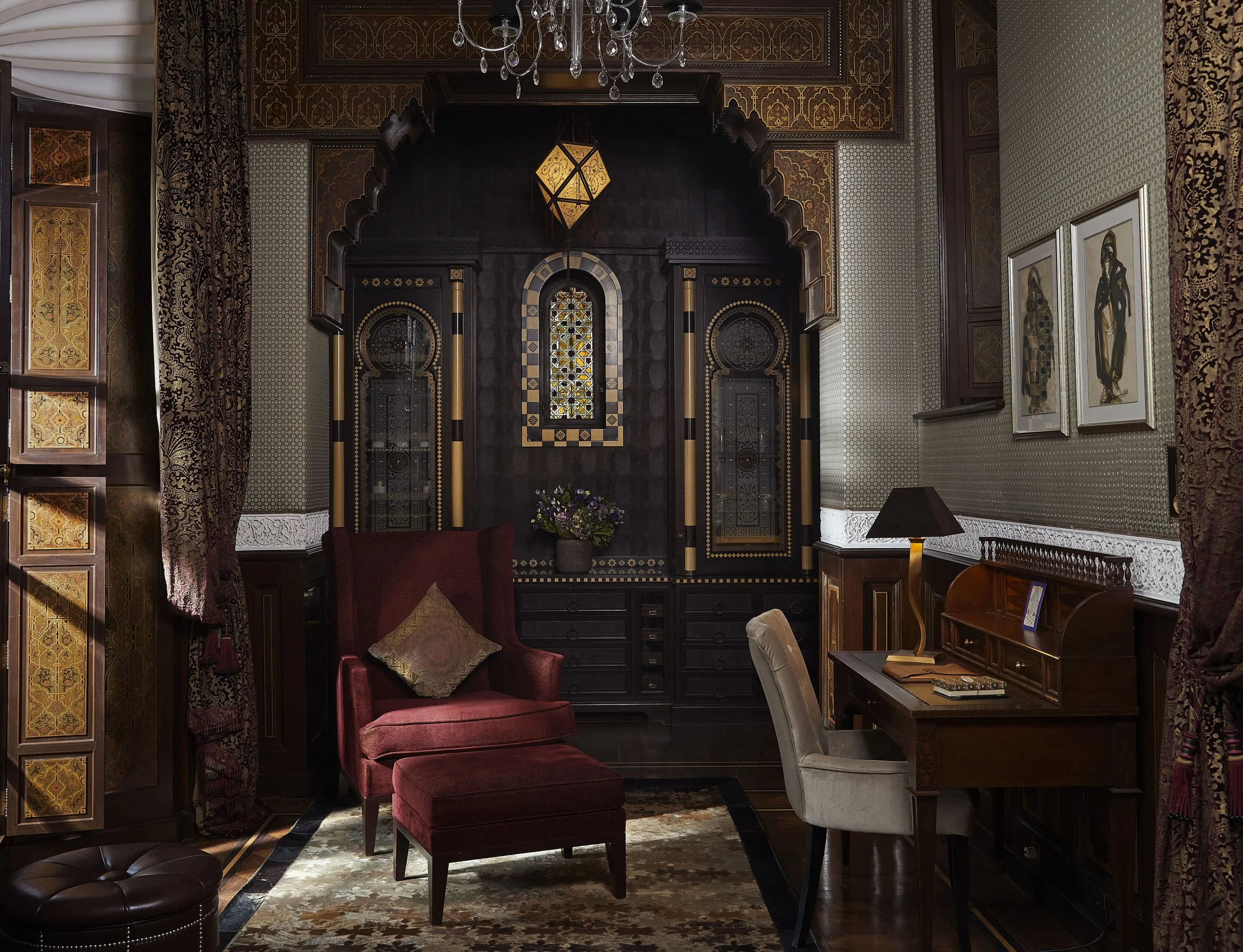 An ornate living room with gold and dark wood decor, a chandelier, framed artwork, a red armchair with decorative pillows, a small table, a dark cabinet with stained glass, and patterned curtains.