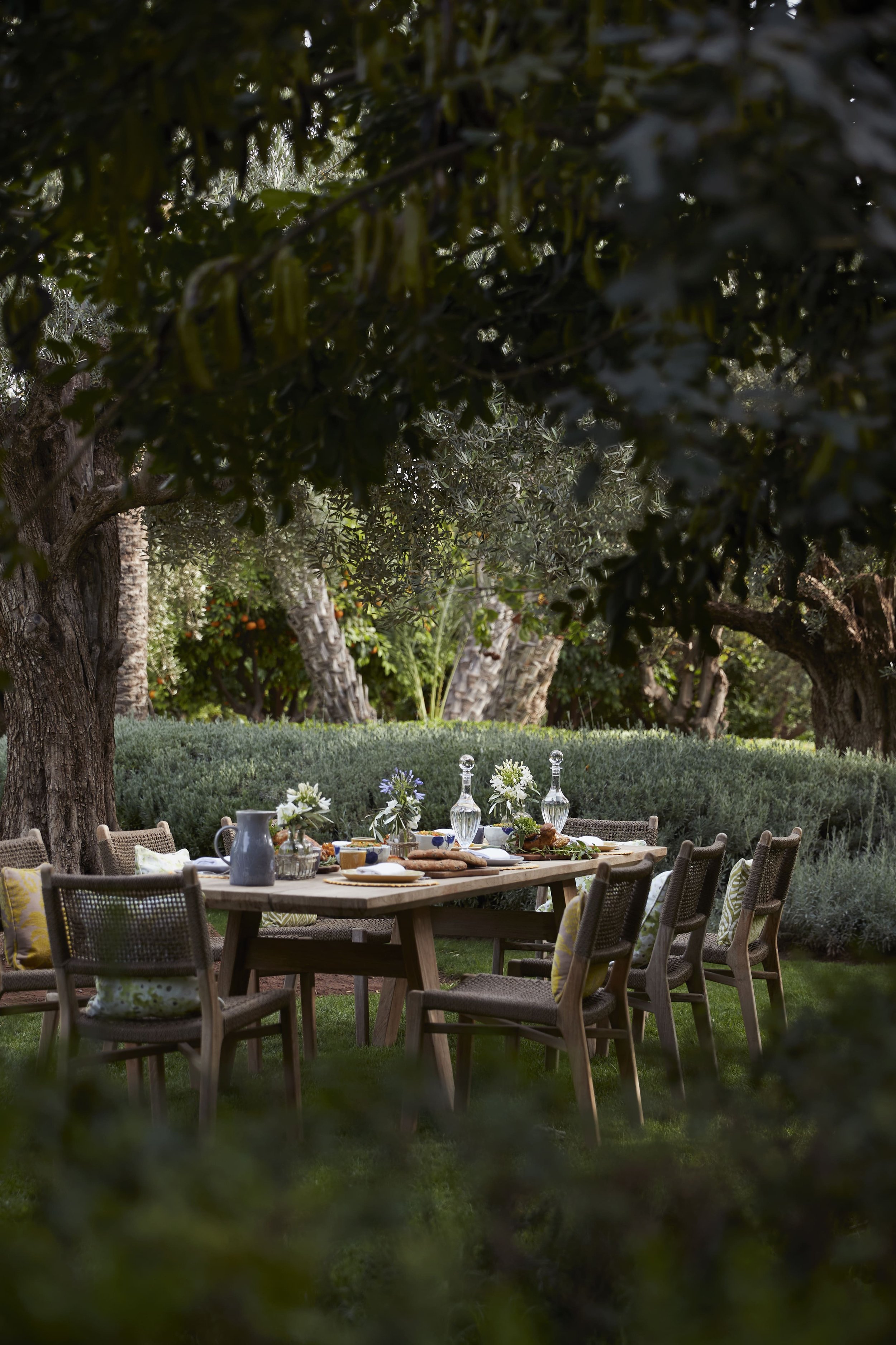 A large outdoor gathering table set for a meal in a lush garden with trees and bushes surrounding it, with various dishes, vases, and glassware.