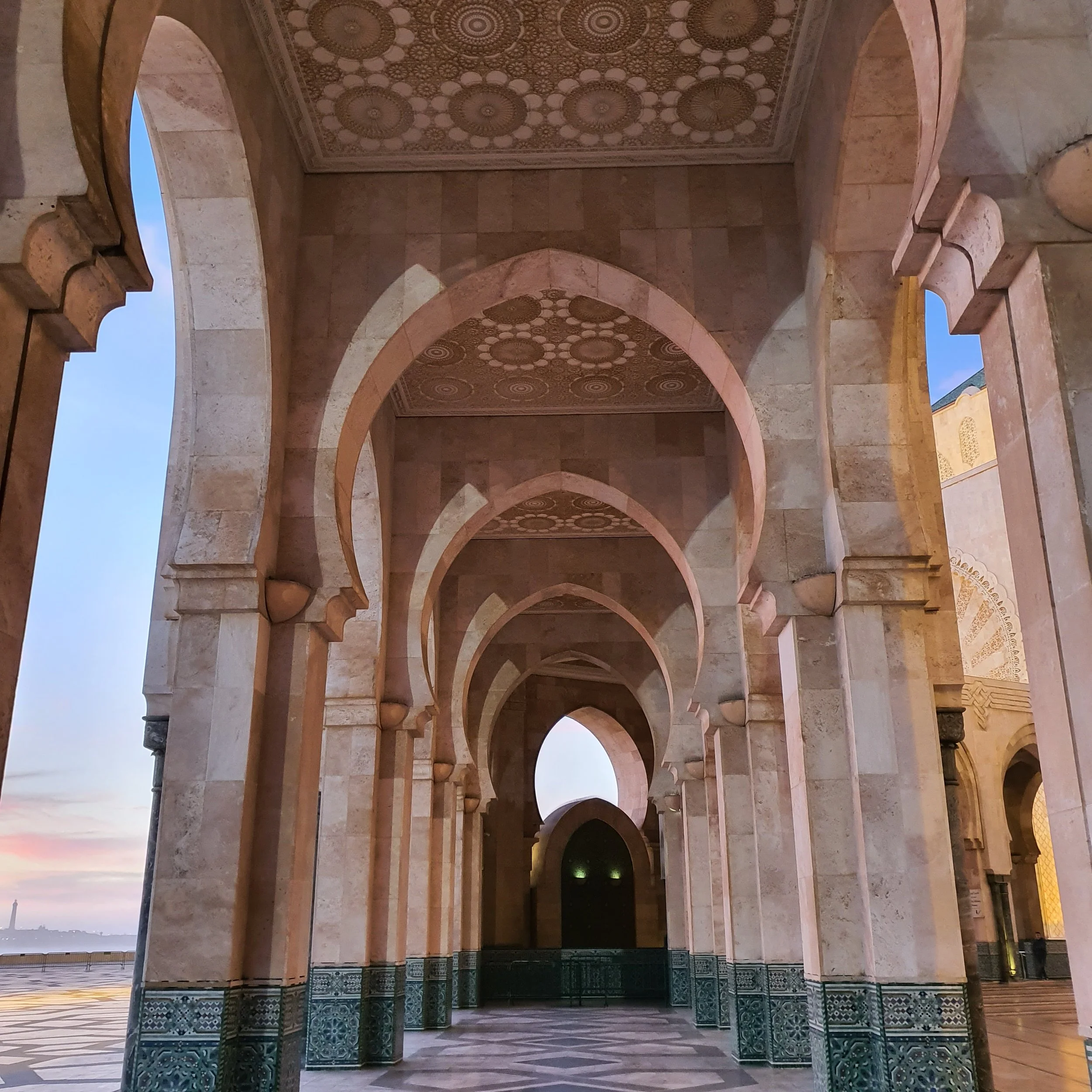 A series of arches in a building with intricate patterned ceilings and tiled columns, viewed from below at sunset.