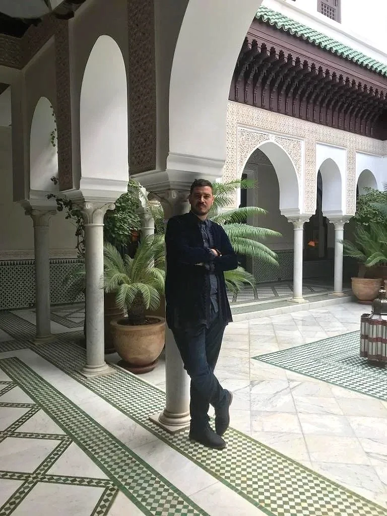 A man in dark clothing standing with arms crossed in a courtyard with arches, columns, potted plants, and tiled floors.