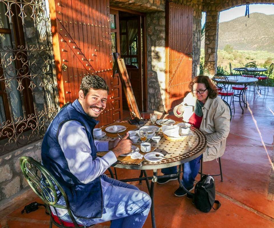 Two people sitting at a round table enjoying coffee and tea, with open wooden gates and scenic mountains in the background.
