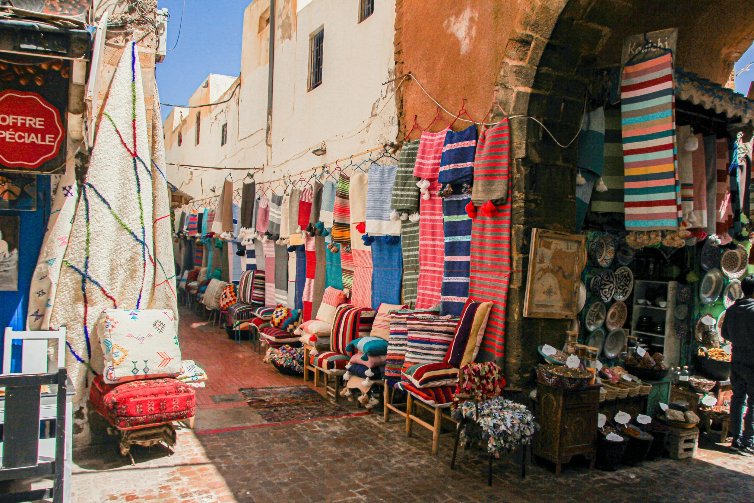 An outdoor market stall displaying colorful textiles, rugs, and handmade crafts on display. The stall is situated on a cobblestone street with buildings in the background, under a bright blue sky.