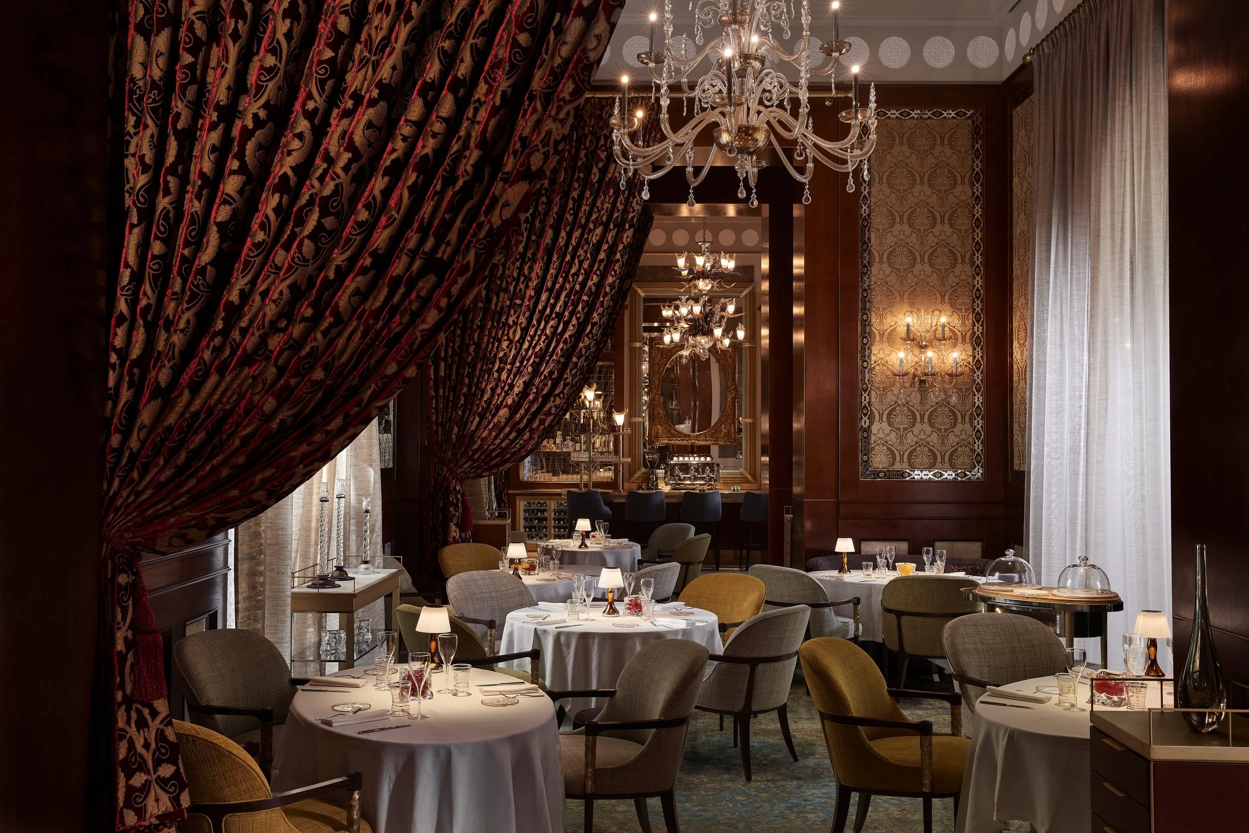 Elegant restaurant dining room with draped red and gold patterned curtains, chandeliers, wall sconces, and tables set with white tablecloths, glassware, and small lamps.