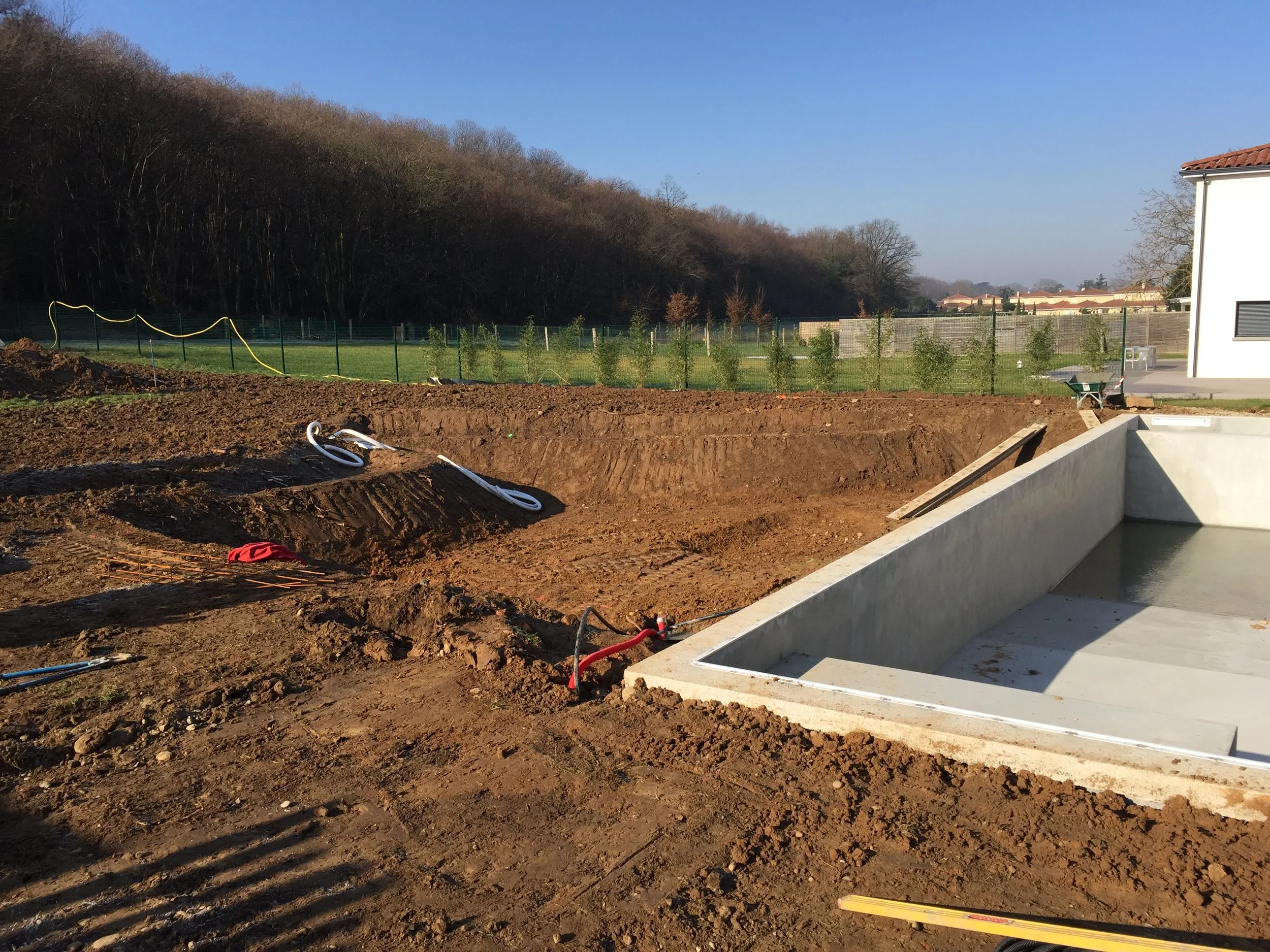 Travaux de construction avec excavation de sol, un bassin en béton en cours de construction, maison moderne à droite, terrain dégagé avec des buissons en arrière-plan et un ciel bleu clair.