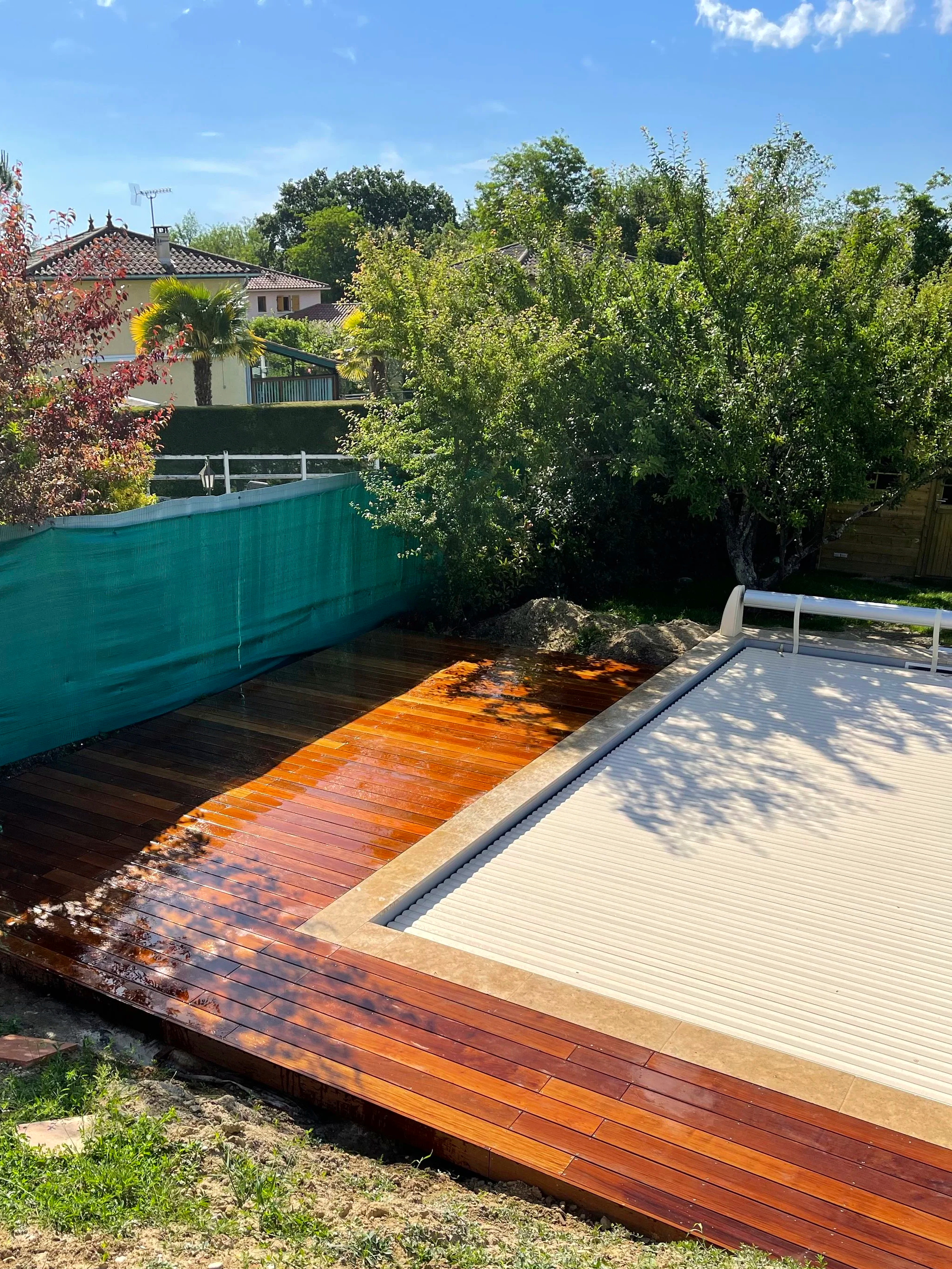 Terrasse en bois rougeâtre avec un espace enroulé, arbres et une maison en arrière-plan, ciel bleu avec quelques nuages.