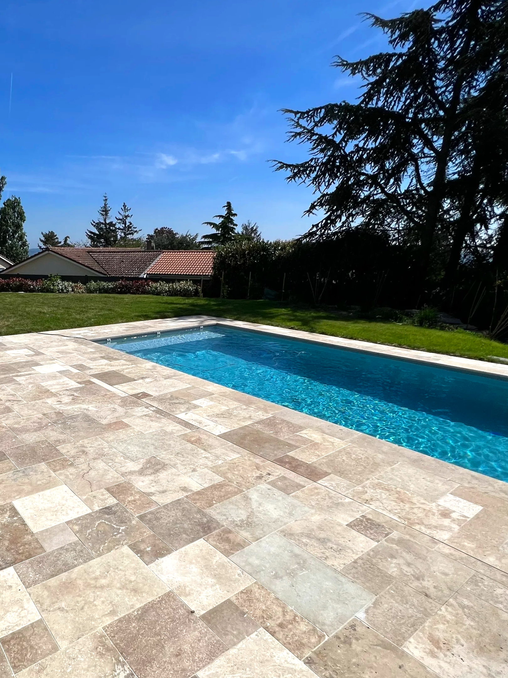 Piscine en pierre entourée d'une terrasse en carreaux beige, grand arbre et hautes haies dans un jardin résidentiel ensoleillé.