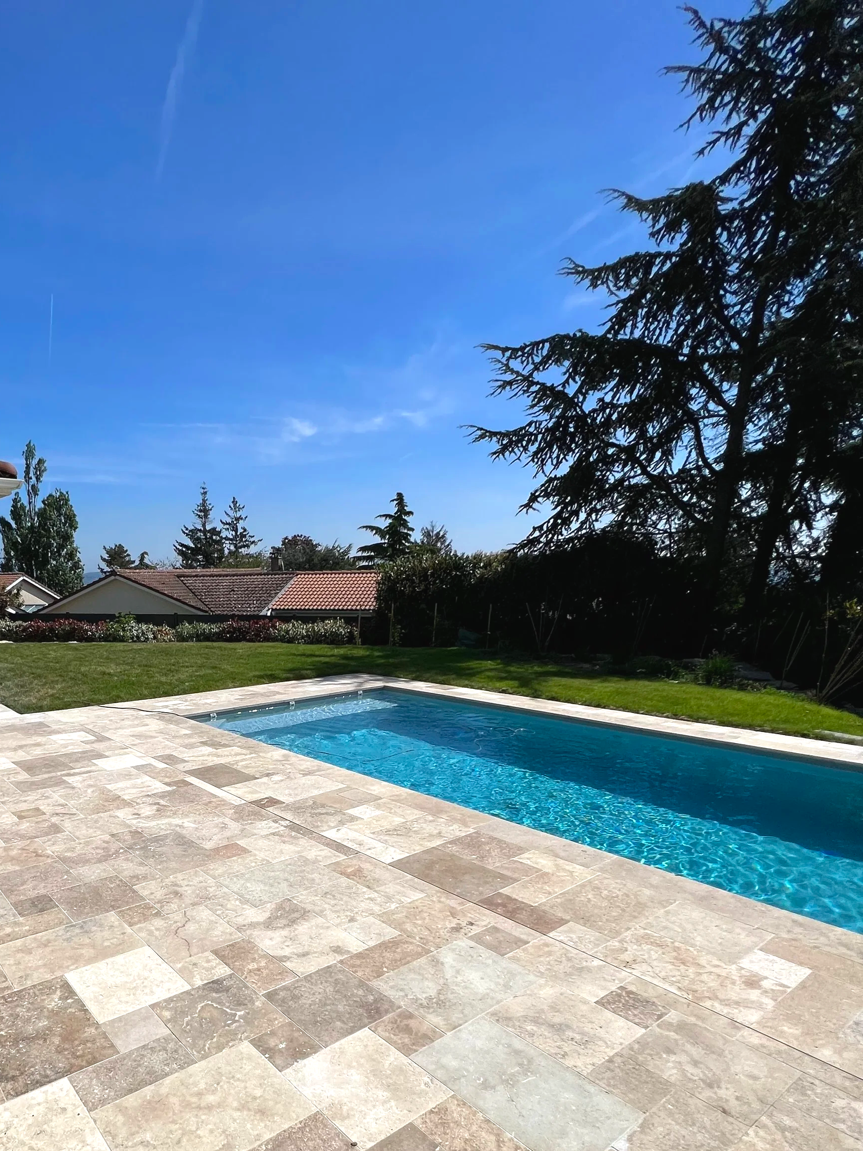 Un piscine en plein air avec un sol en pierre, entourée de verdure, des maisons en arrière-plan, et un ciel bleu avec quelques nuages.