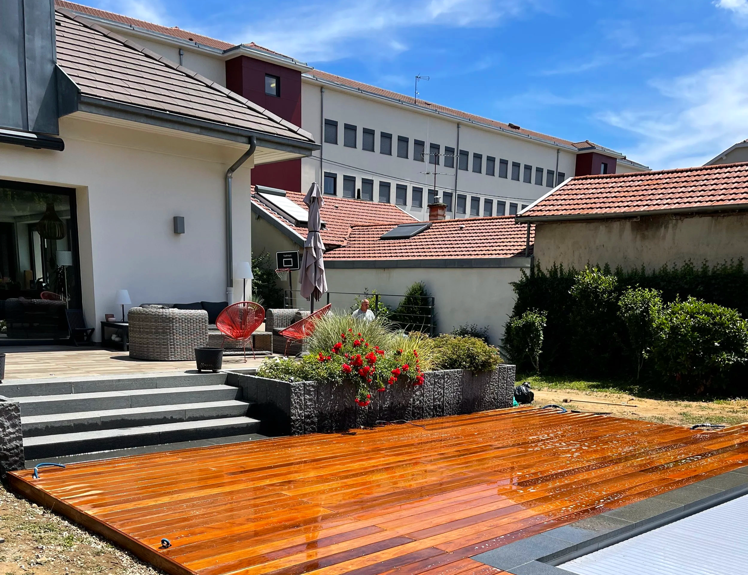 Ciel bleu au-dessus d'un immeuble résidentiel avec une terrasse en bois fraîchement rénovée, des meubles d'extérieur, des plantes, une piscine et une maison avec toit en tuiles.