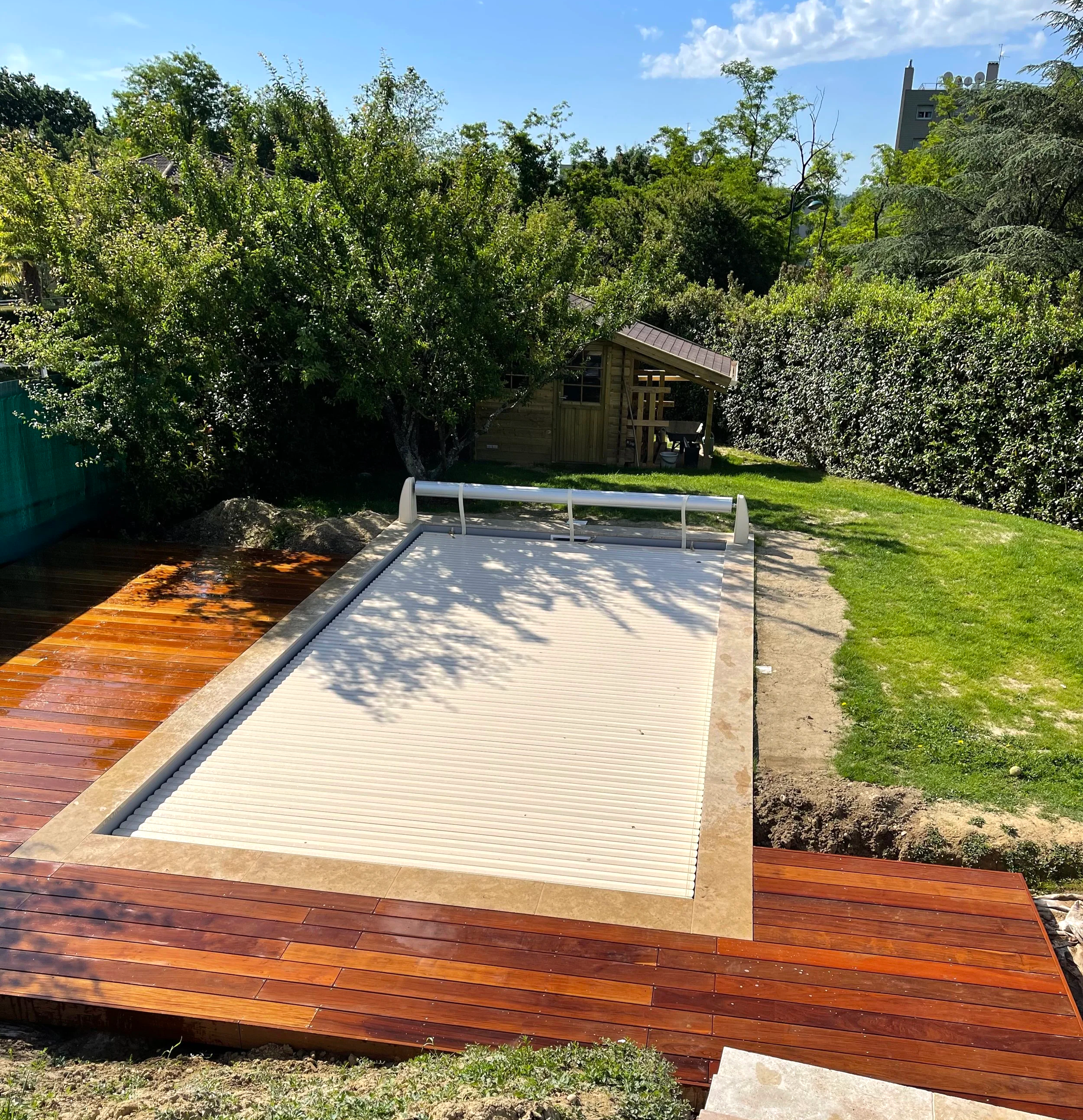 Une installation de nouvelles terrasses en bois, avec une section en bois non traité et une section en bois traité, dans un jardin avec arbres et une petite maison en arrière-plan.