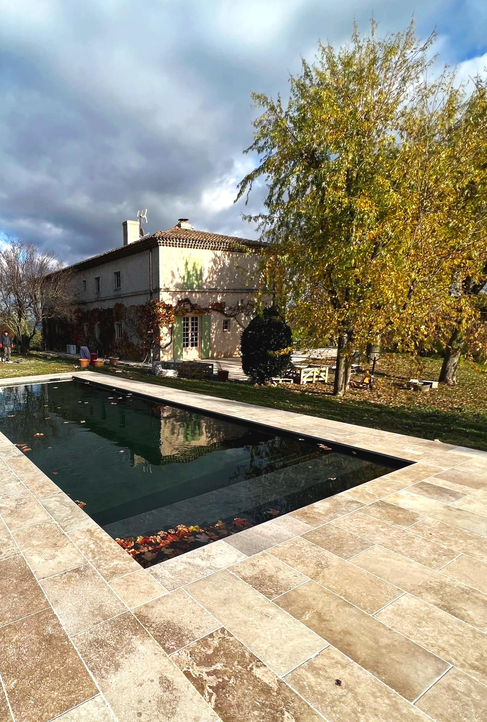 Maison ancienne avec cheminée, volets verts et volets fermés, entourée d'arbres en automne avec feuilles dorées, reflet dans la piscine. Ciel nuageux.