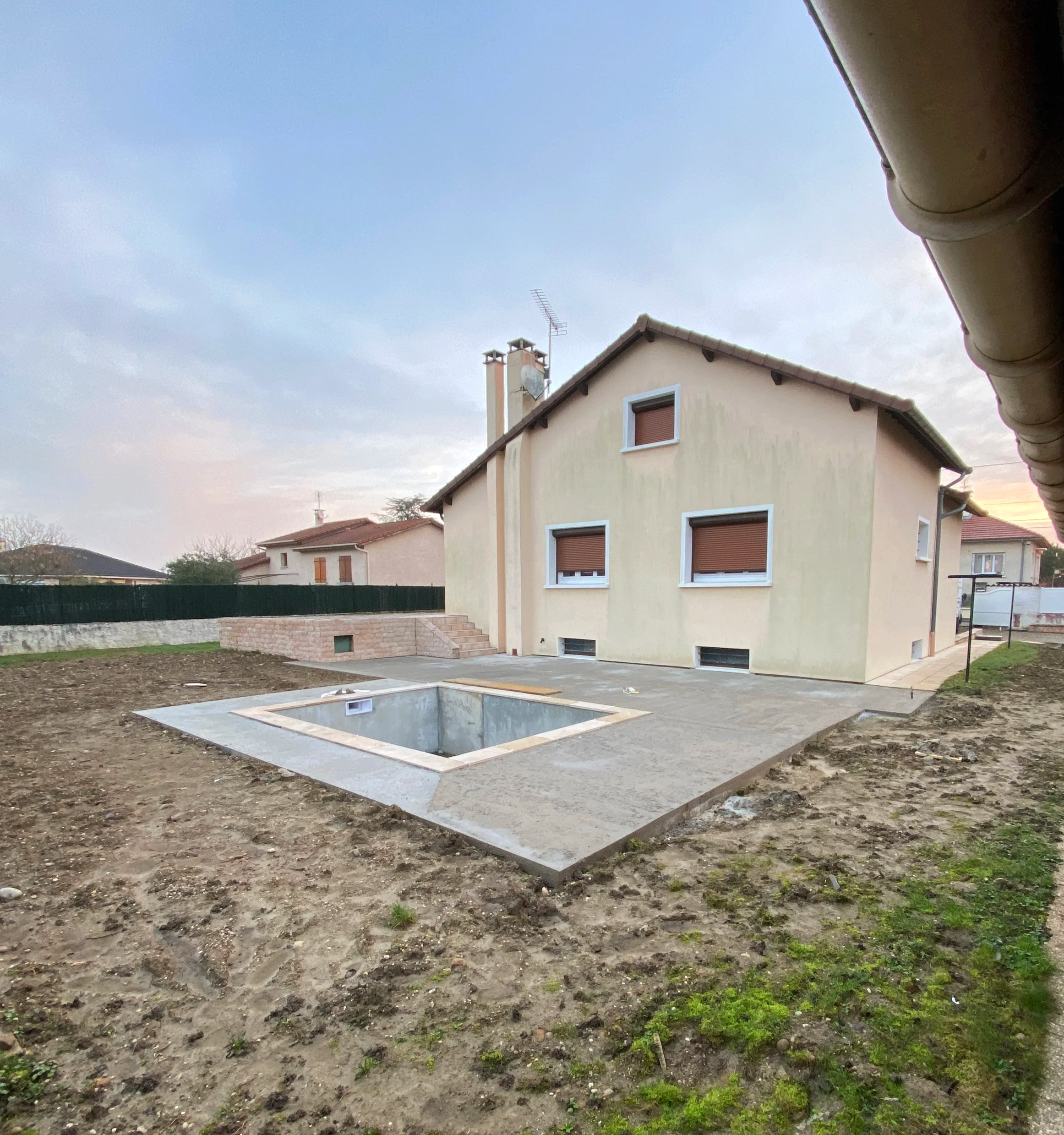 Un terrain en cours de préparation avec une excavatrice creusant un trou pour une piscine hors sol en béton, une maison avec des fenêtres à volets roulants, un ciel nuageux et une pelouse entre les zones de construction.