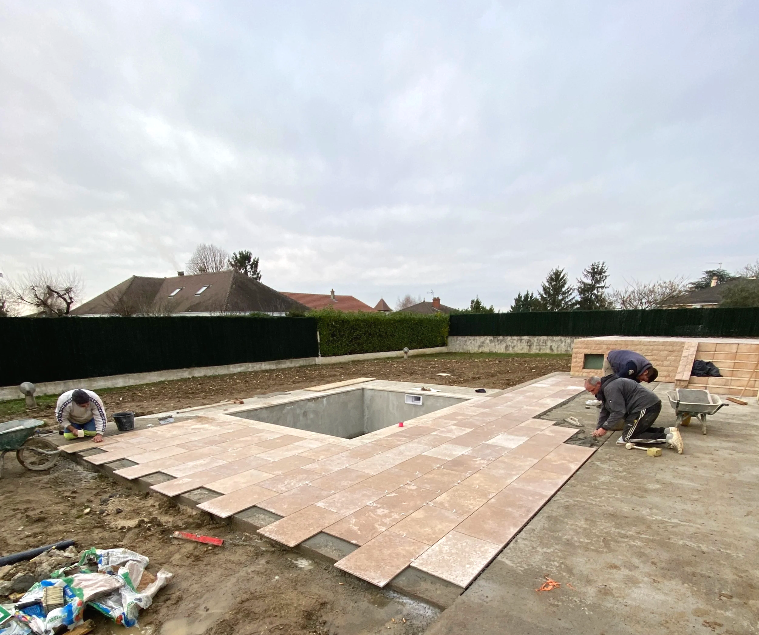 Travaux de pose de pavés en extérieur par deux personnes, avec un jardin et des maisons en arrièree-plan.