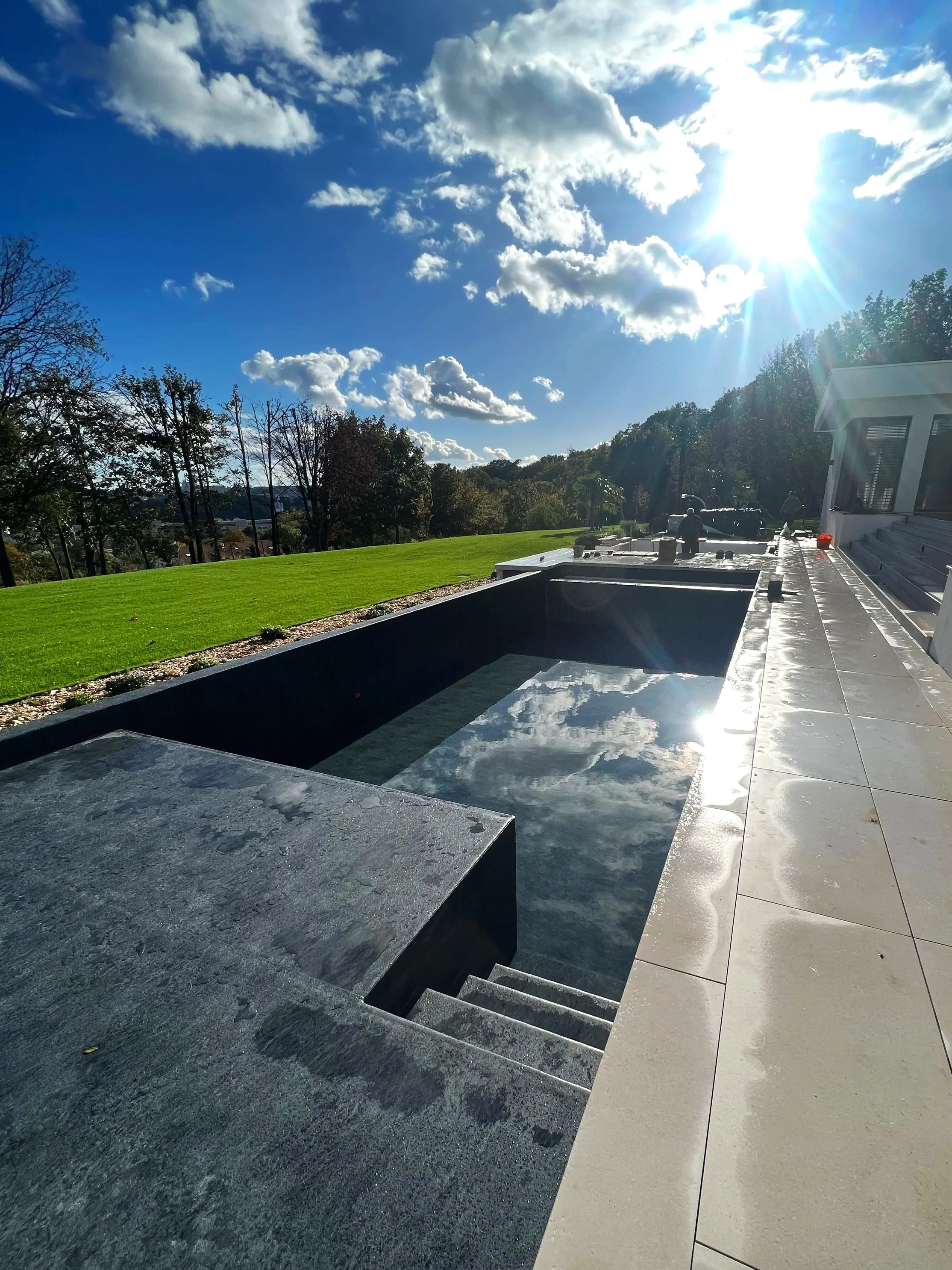 Une piscine noire en béton avec de l'eau très claire, entourée d'une terrasse en carrelage blanc, avec un bâtiment moderne en arrière-plan et une vue sur un ciel ensoleillé avec quelques nuages.