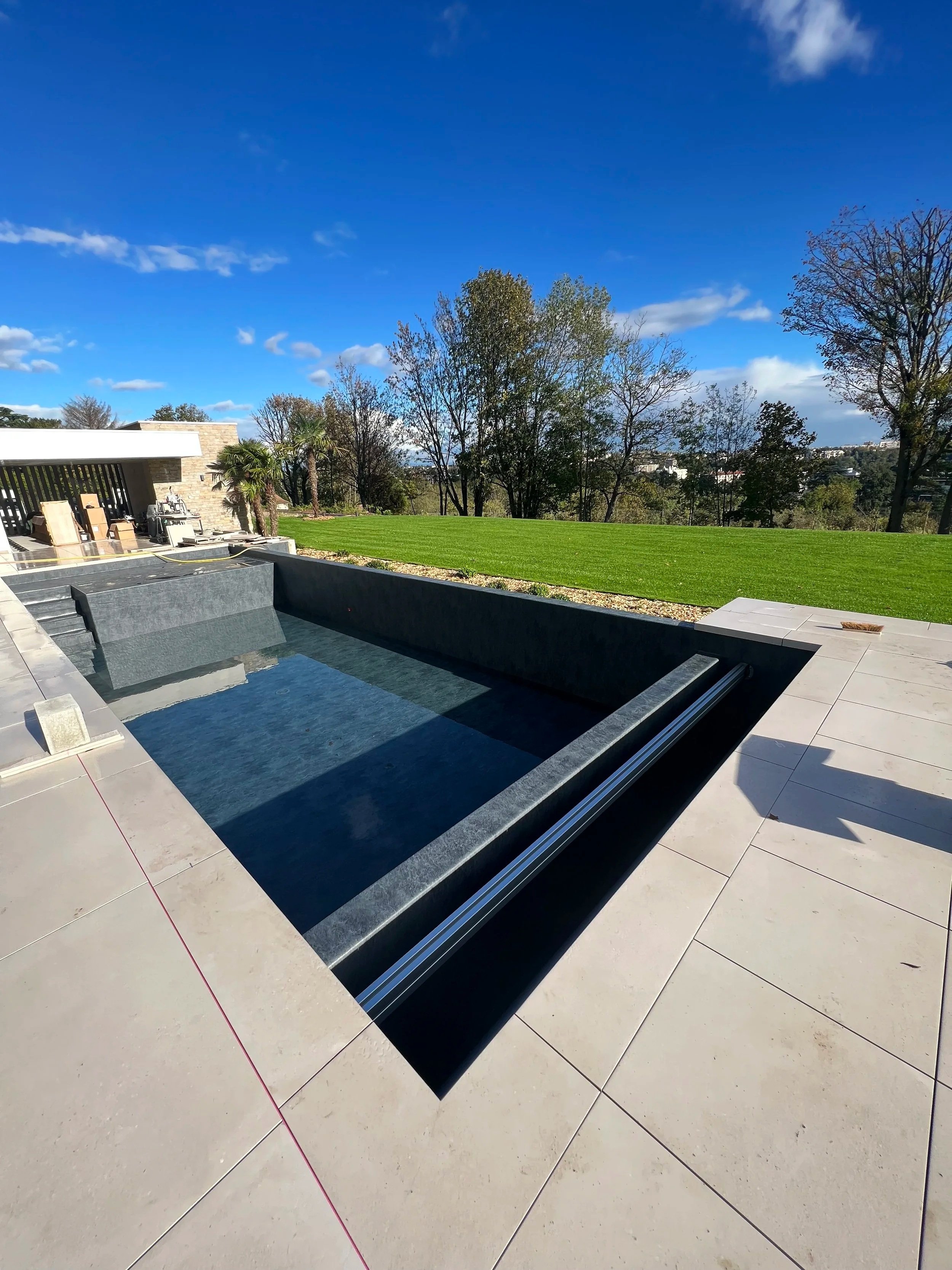 Vue d'une piscine moderne avec un bord en pierre noire, une zone dallée en beige, un jardin bien entretenu avec pelouse verte et arbres, sous un ciel bleu avec quelques nuages.