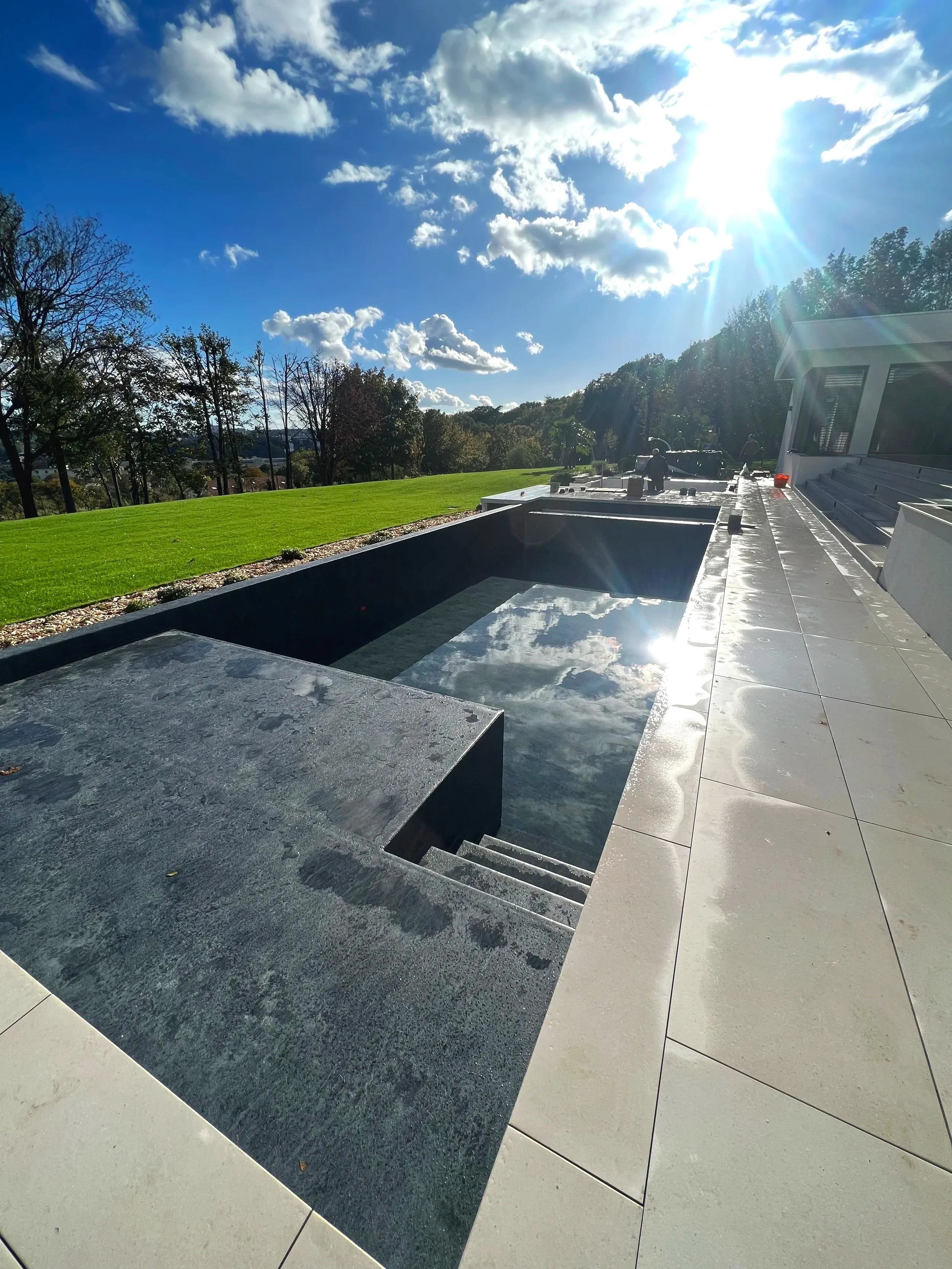 Une terrasse moderne avec bassin d'eau, sols en pierre, vue sur un ciel ensoleillé avec des nuages, pelouse verte et arbres en arrière-plan, bâtiment blanc à droite.