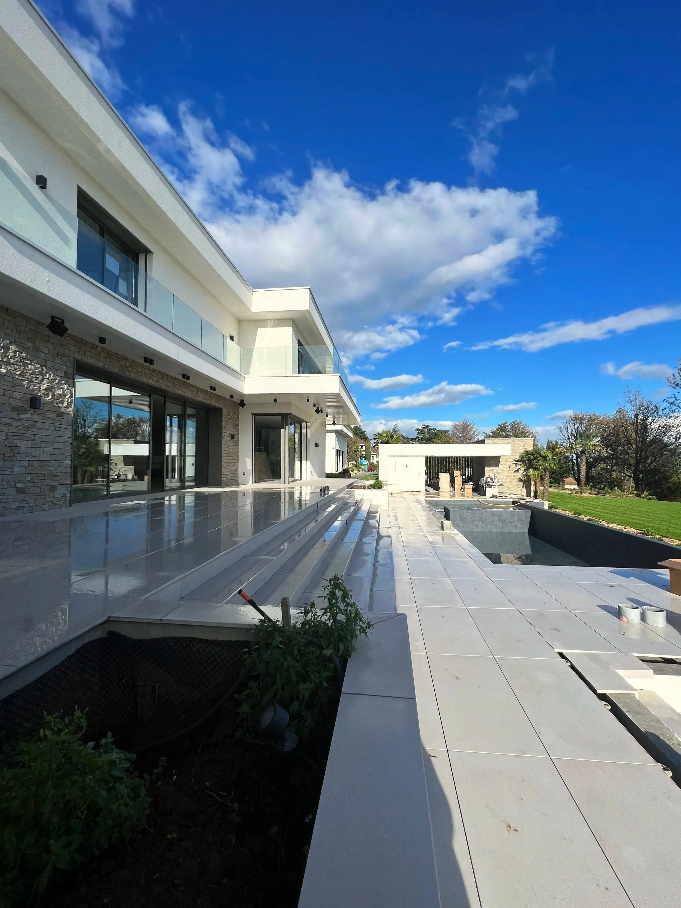 Une maison moderne en construction avec de grandes fenêtres, un patio en pierre et un ciel bleu avec des nuages blancs.