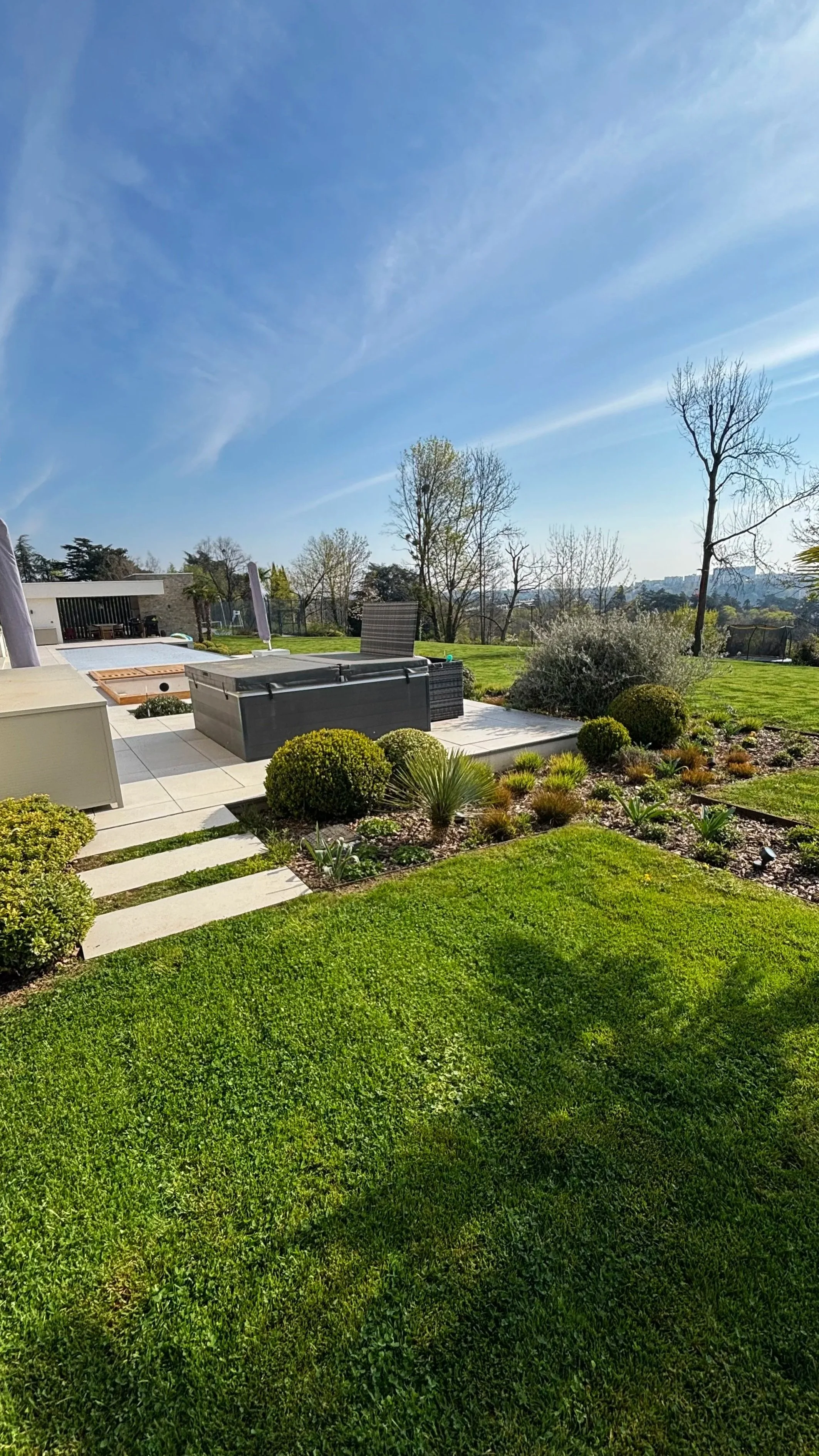 Jardin bien entretenu avec pelouse verte, buissons, arbres et espace détente avec mobilier.