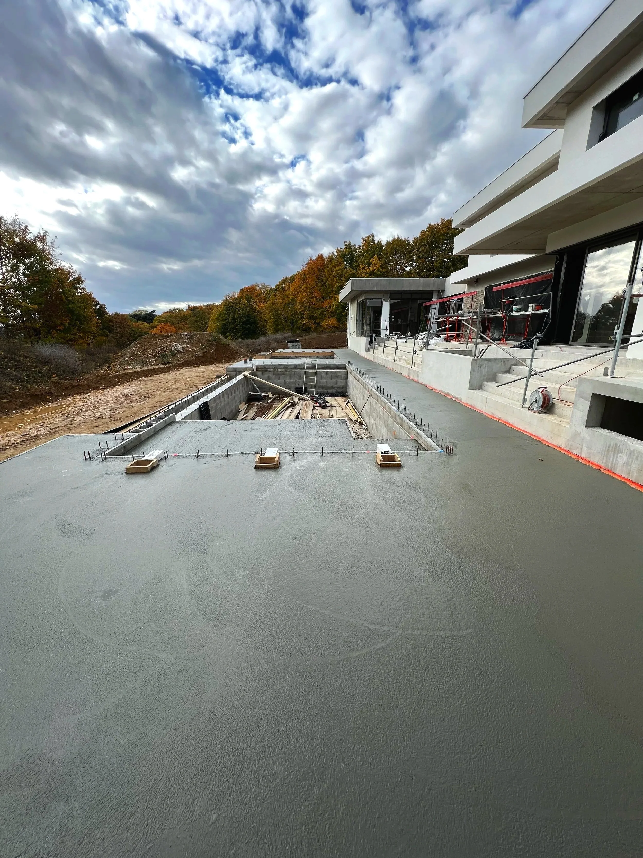 Construction d'une piscine en béton à côté d'un bâtiment moderne, avec un ciel nuageux en arrière-plan.
