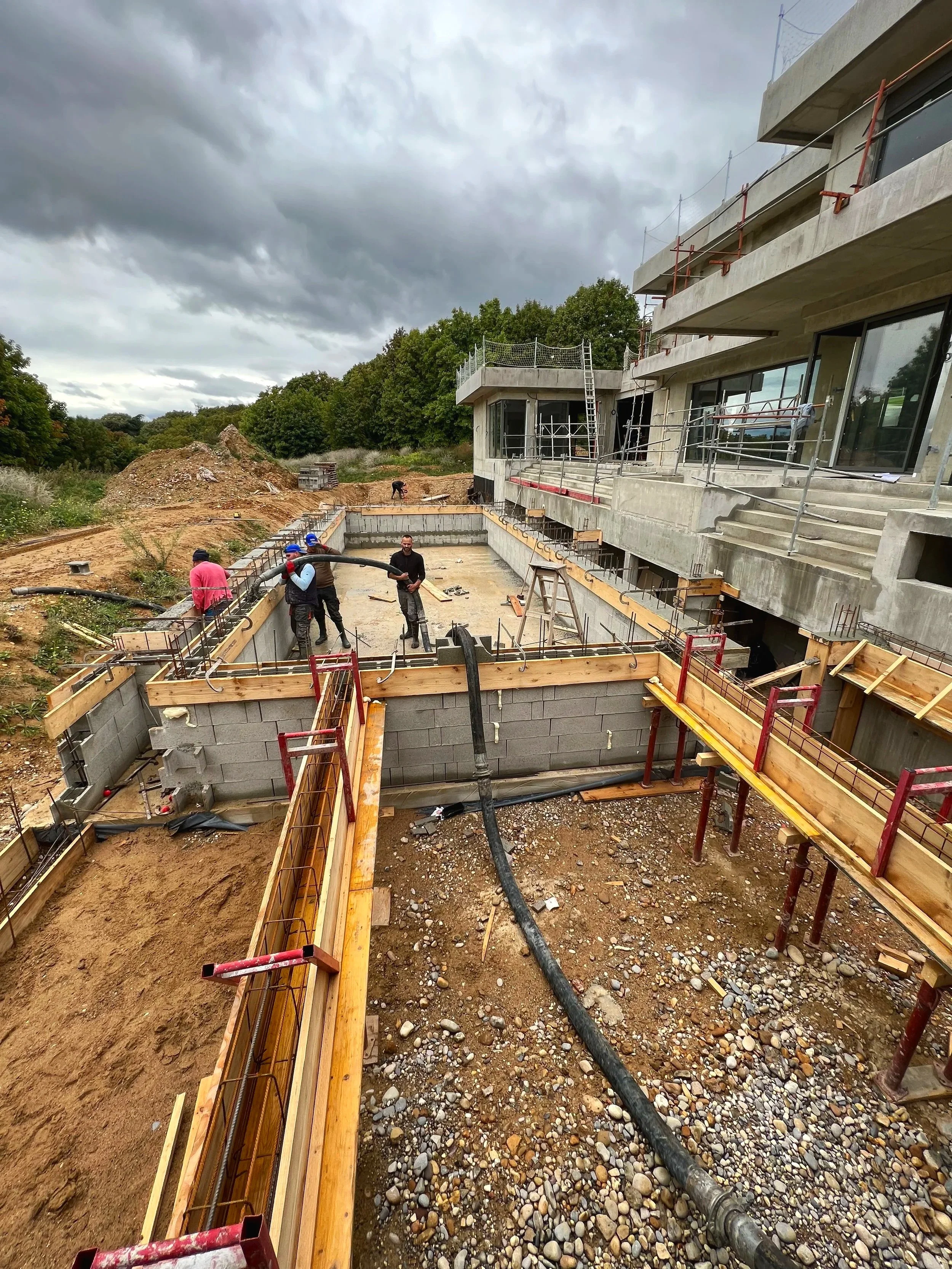 Construction d'une piscine attenante à une maison moderne avec une terrasse en béton et des barrières de sécurité, plusieurs ouvriers travaillent, le ciel est nuageux.