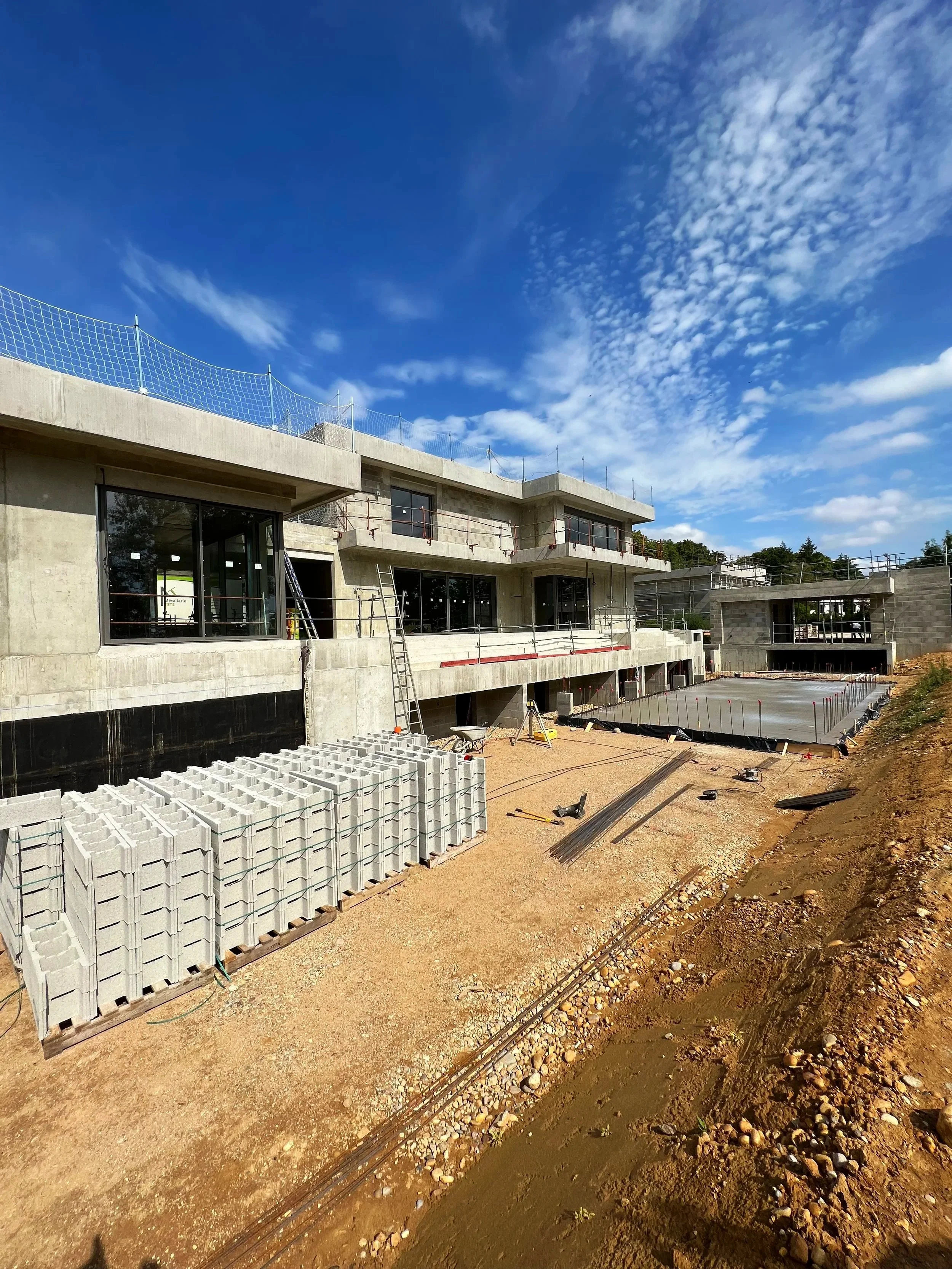 Construction d'un bâtiment moderne avec structure en béton, fenêtres et murs en cours de finition. Apparentement, un espace extérieur en préparation avec des matériaux de construction et des outils. Ciel bleu avec quelques nuages.