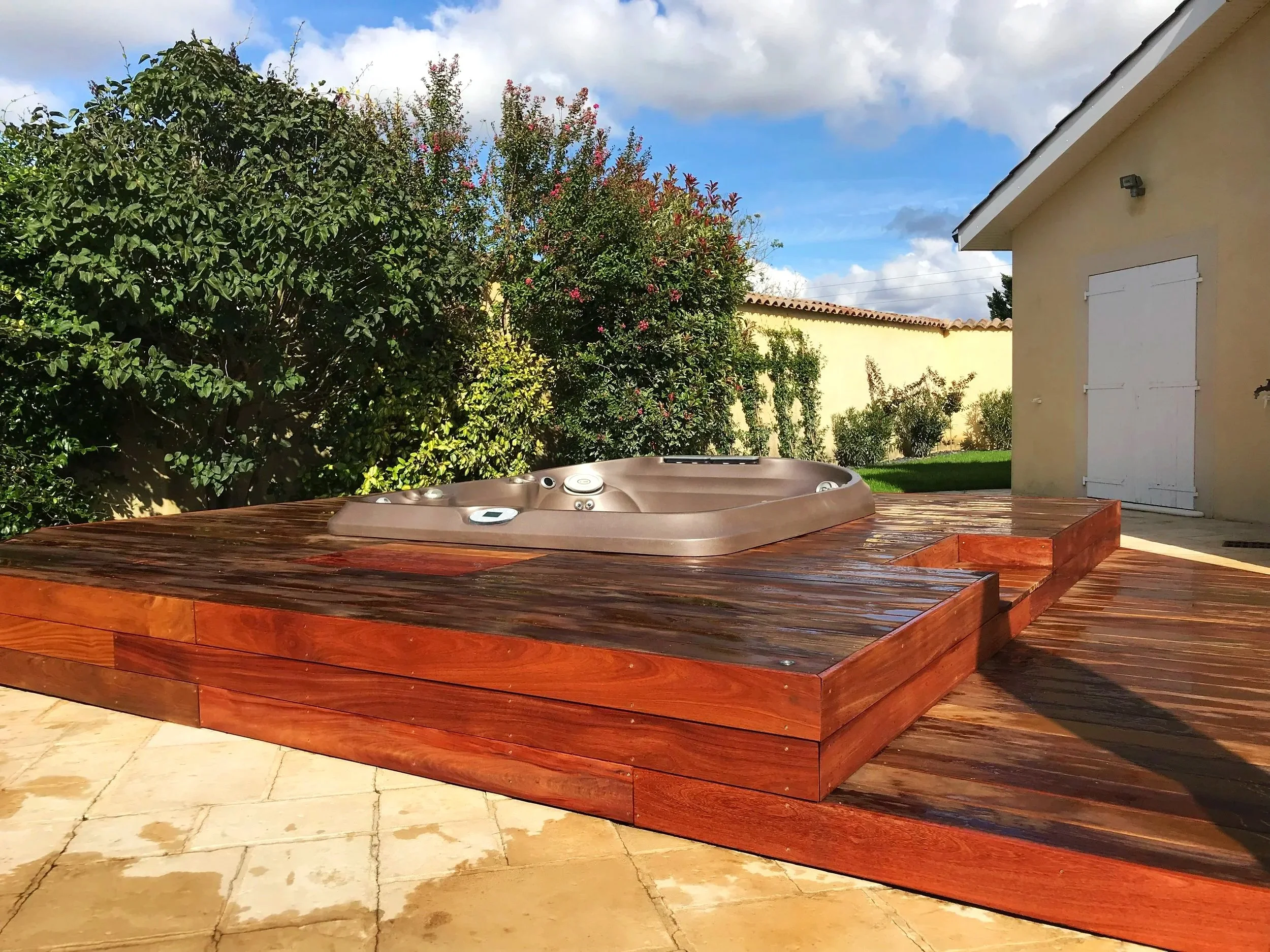 Un jacuzzi installé sur une plateforme en bois en extérieur, entouré de végétation avec des arbres et un mur. Le ciel est bleu avec quelques nuages.
