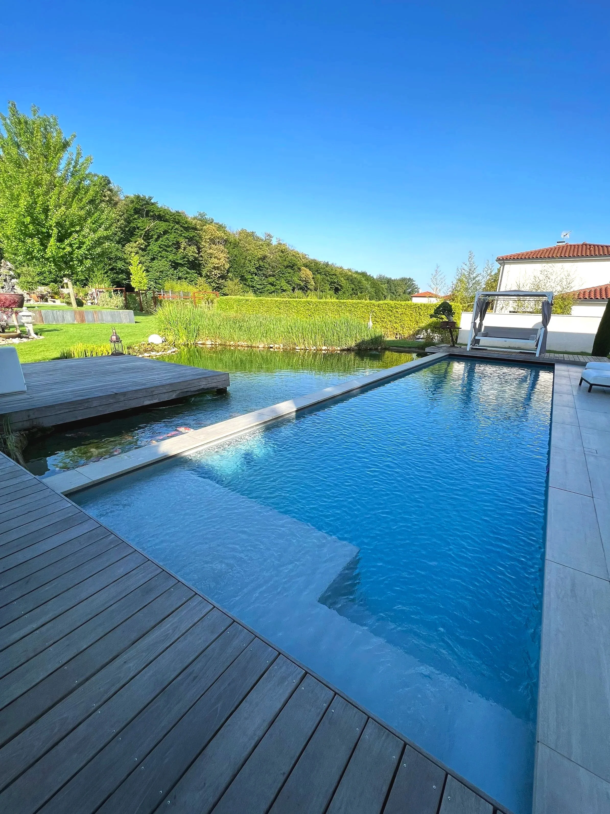 Piscine extérieure en bois avec un espace détente, entourée de verdure et d'une maison avec un toit en tuiles rouges, sous un ciel bleu clair.