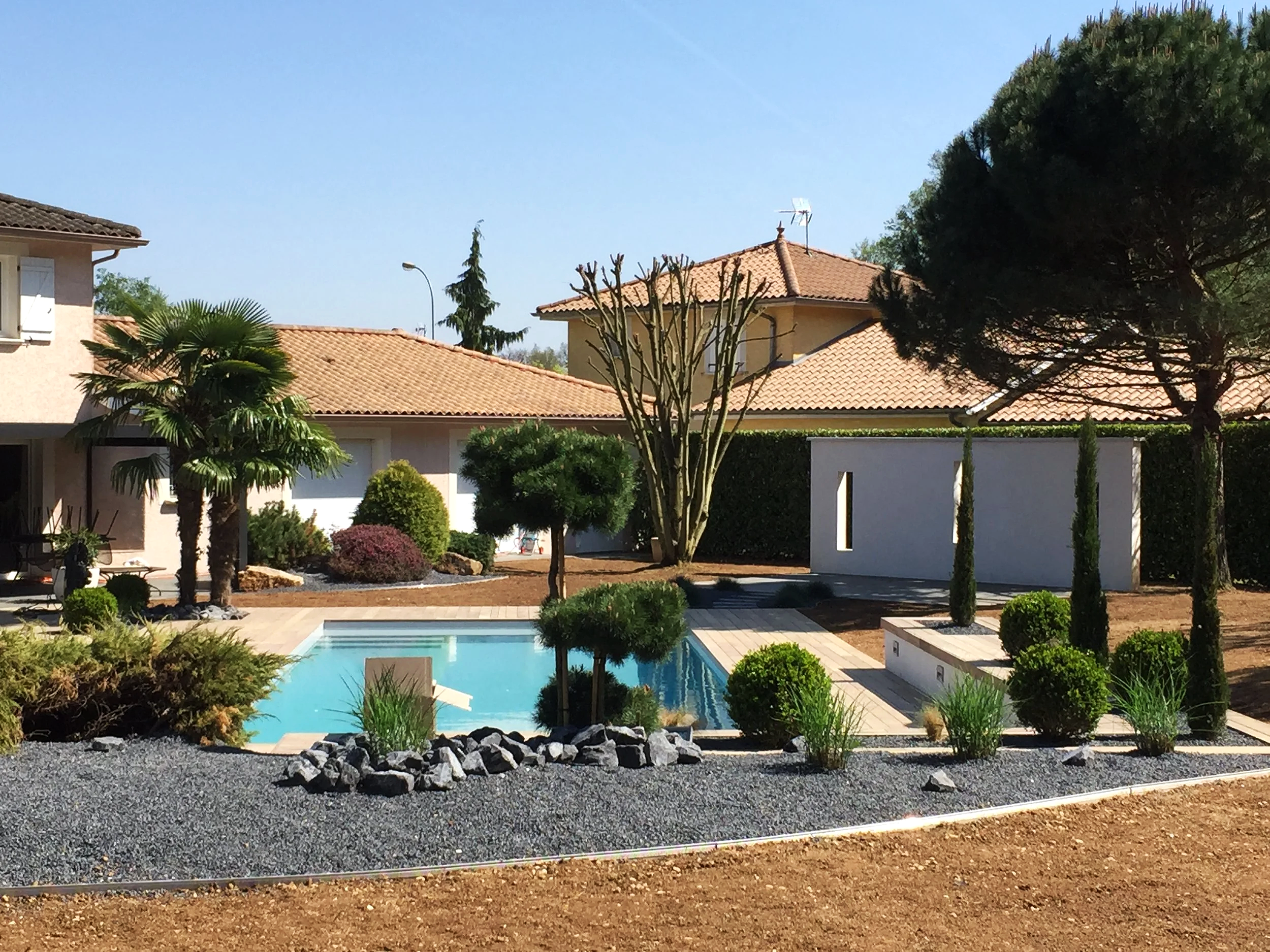 Photo d'une maison avec un toit en tuiles orange, un jardin bien entretenu avec de nombreux arbres et arbustes, une piscine et une allée en pavés. Le ciel est clair et ensoleillé.