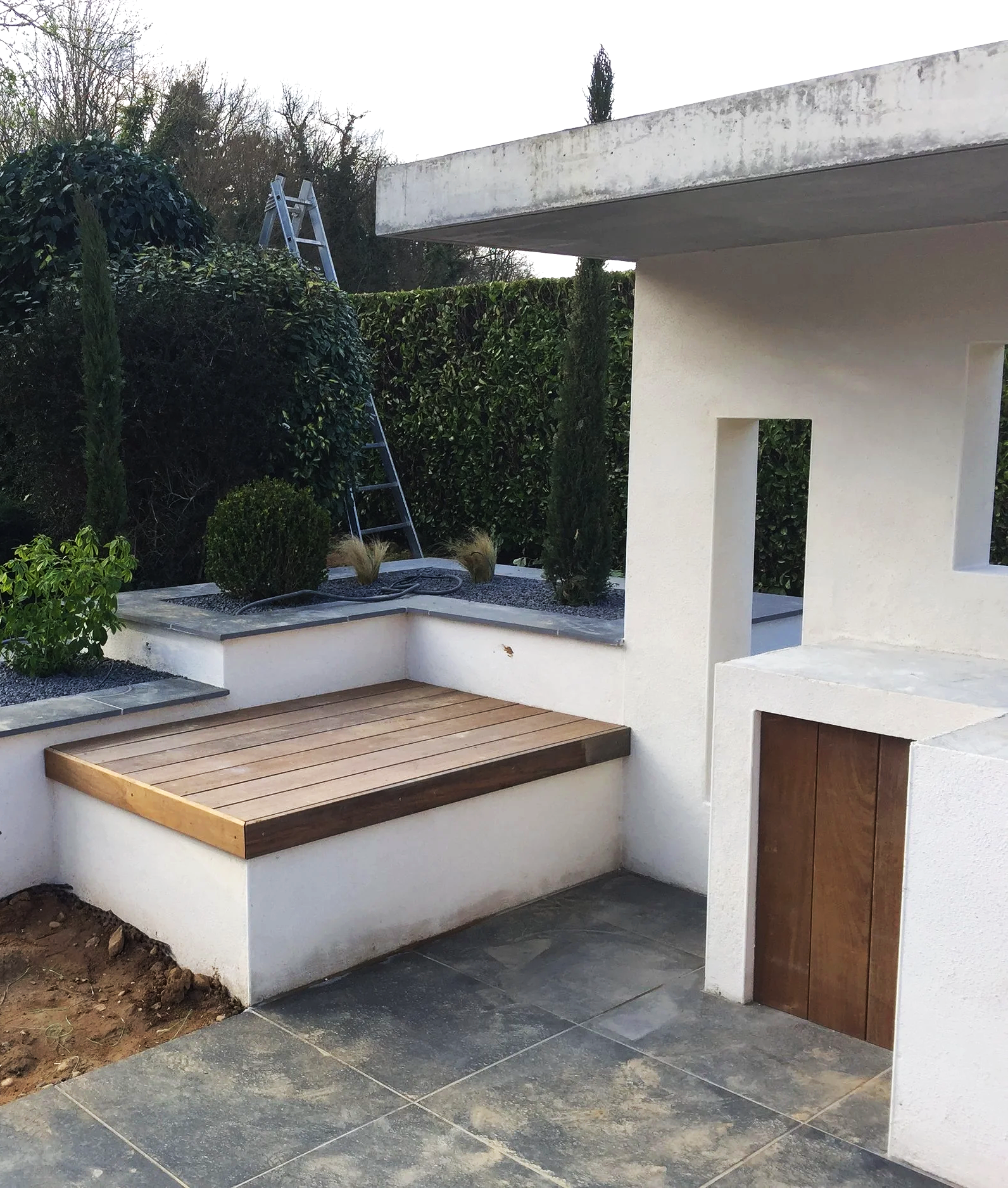 Un espace extérieur avec une structure en béton blanc, une terrasse en bois, des plantes en pot, un jardin bien entretenu avec des haies de buissons, et une échelle en métal contre la haie.
