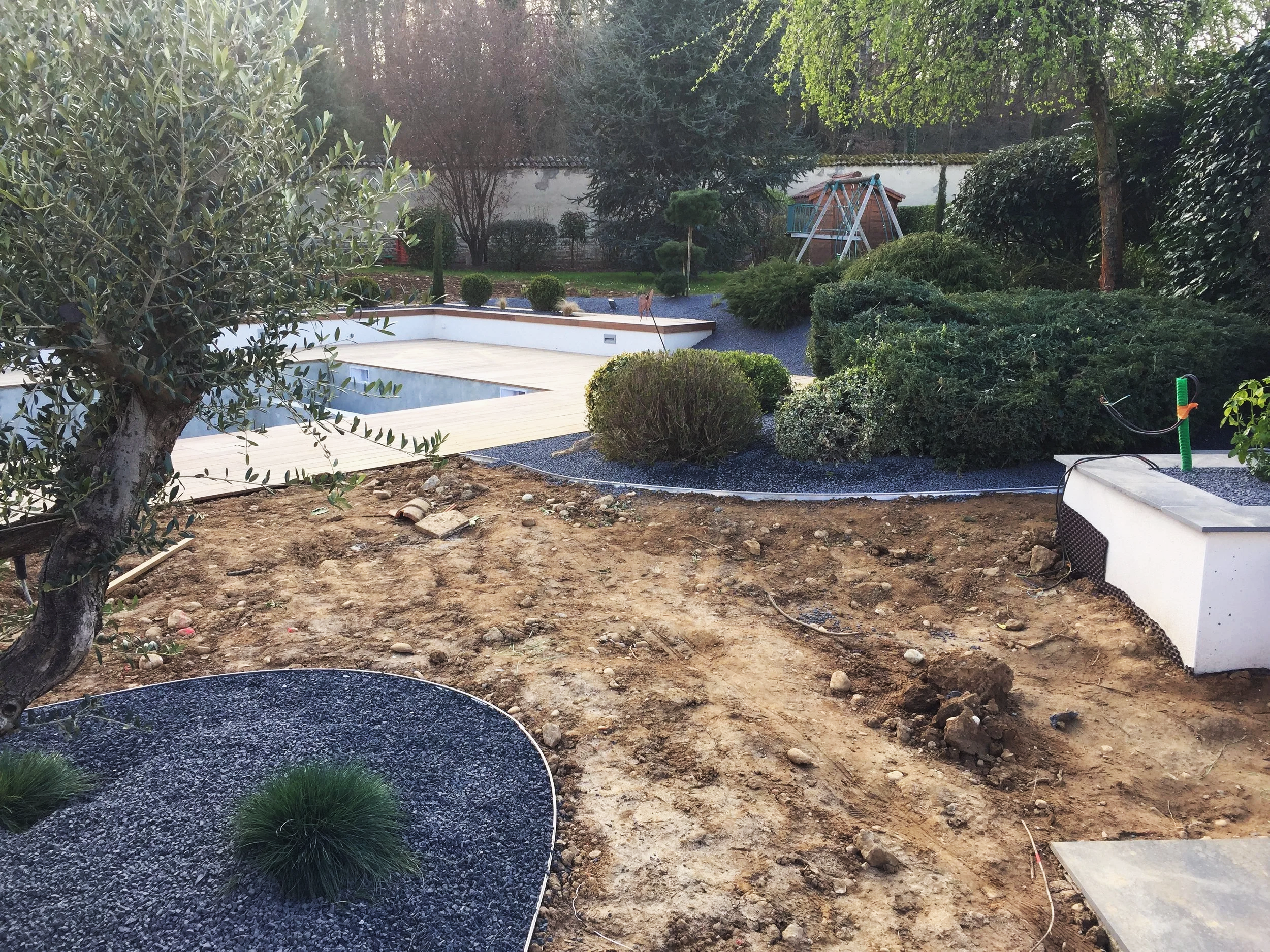 Jardin en cours d'aménagement avec des plantes, un espace de jeux pour enfants, et un sol en construction avec quelques pierres visibles.
