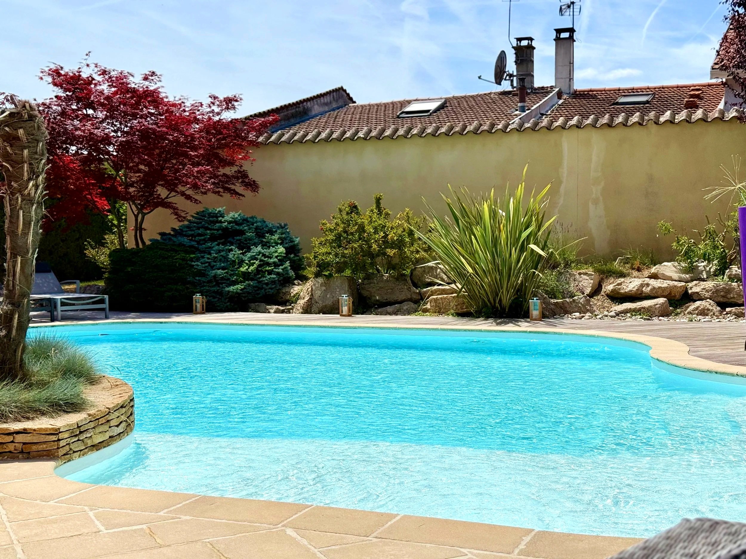 Piscine extérieure avec eaux bleues claires, entourée de dalles en pierre et de végétation variée, avec une maison en arrière-plan, toit en tuiles et arbres, sous un ciel bleu.