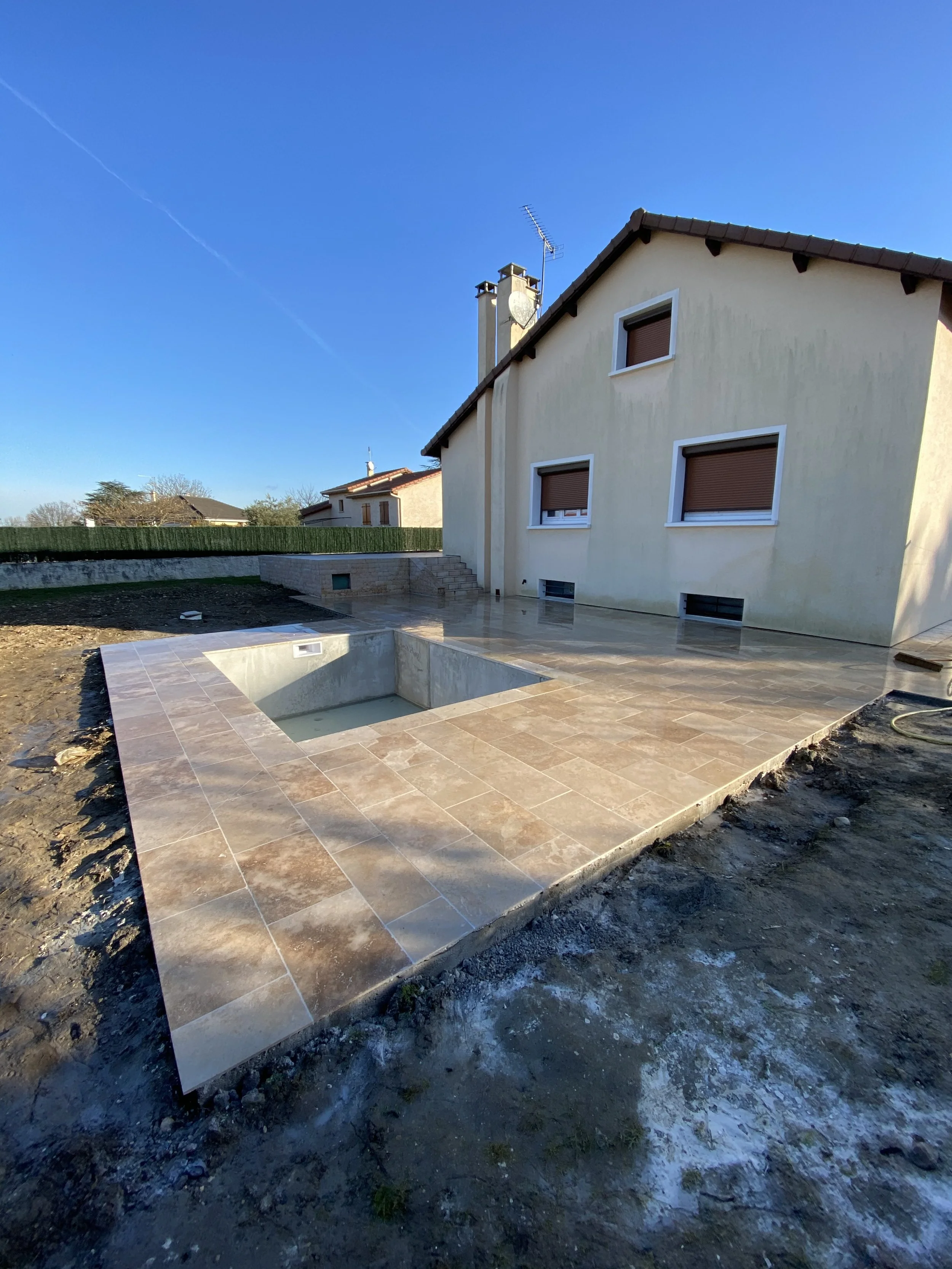 Une maison avec un jardin en construction, terrasse en carrelage beige, terrain non aménagé, ciel bleu clair.