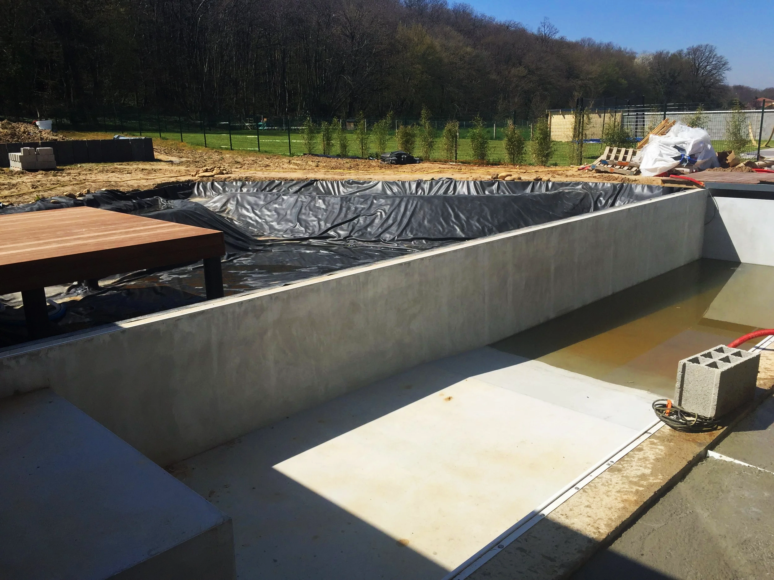 Construction d'une piscine située dans un jardin, avec un bassin en cours de réalisation et des matériaux de construction autour.
