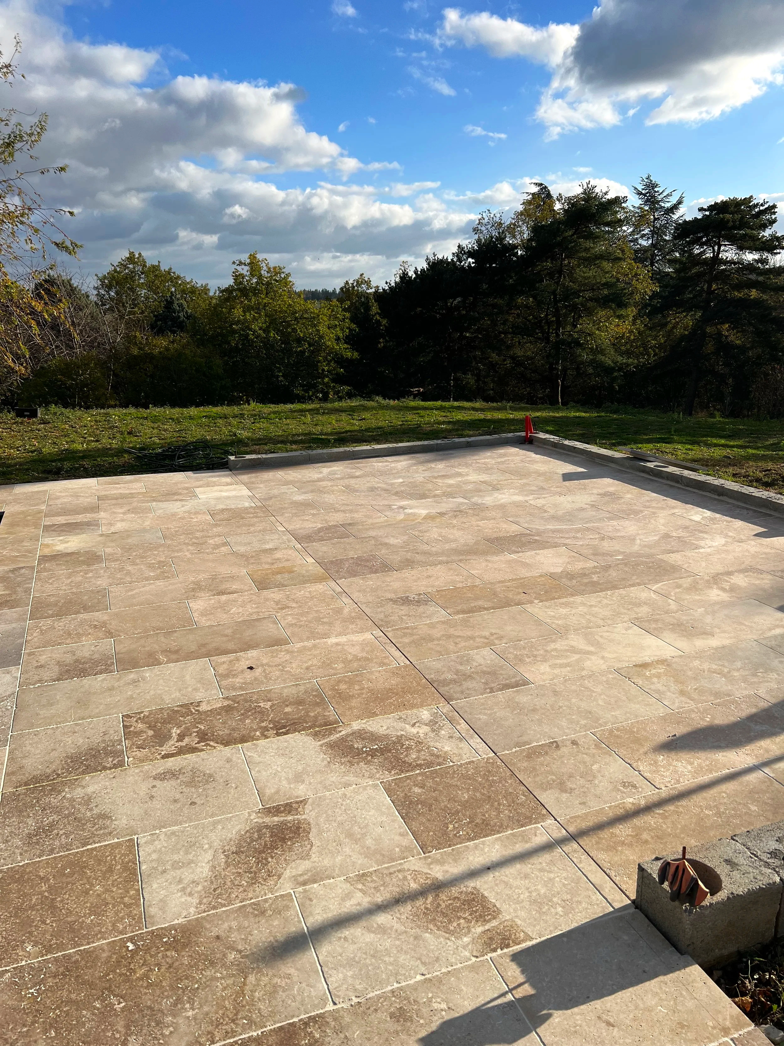 Vue d'une terrasse pavée en pierre avec des plans d'herbe et des arbres en arrière-plan sous un ciel partiellement nuageux.