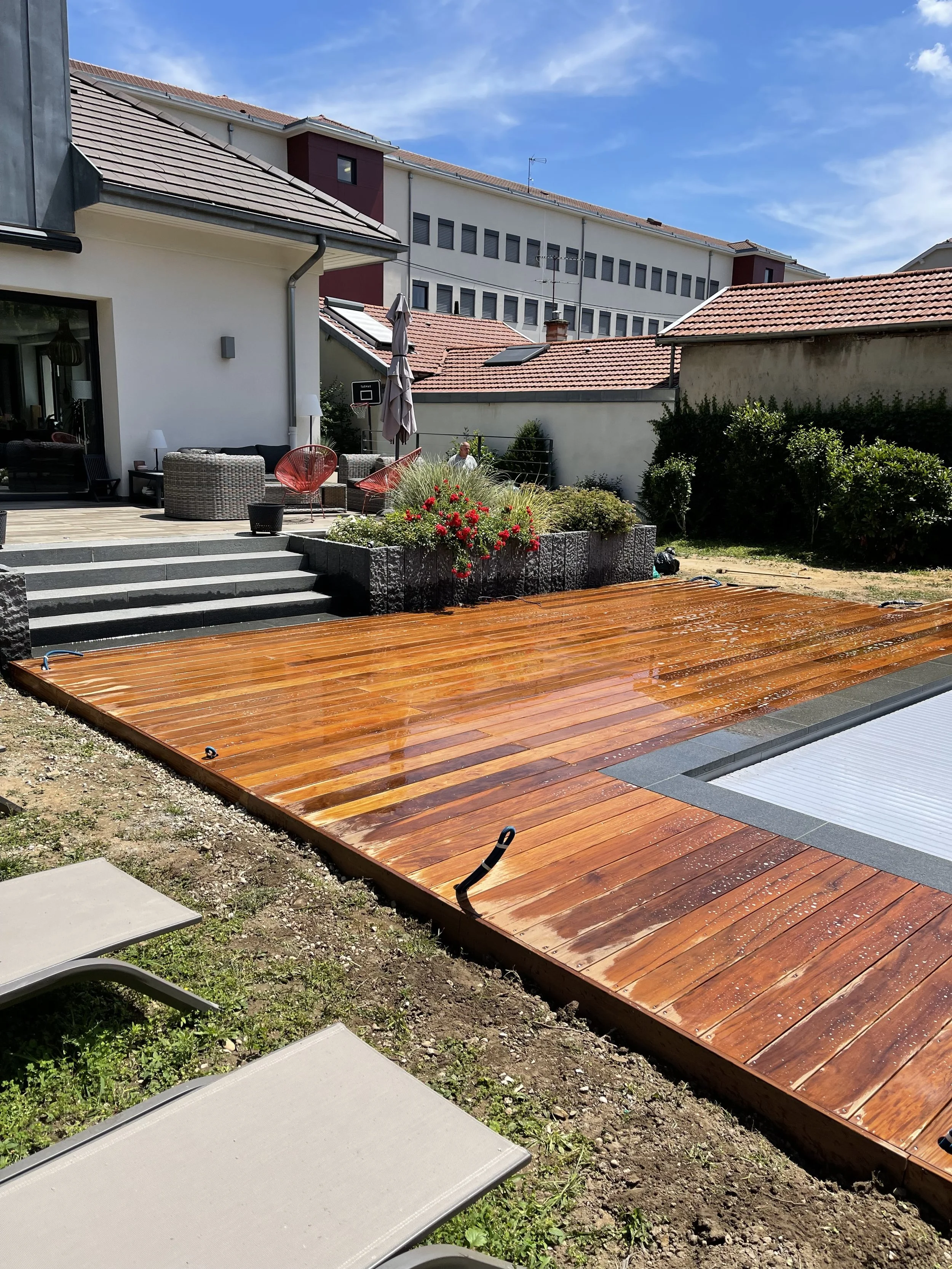 Un patio résidentiel avec une terrasse en bois fraîchement posée, des meubles de jardin, des plantes en fleurs, un parasol, et des voisins discutant sous un ciel ensoleillé.
