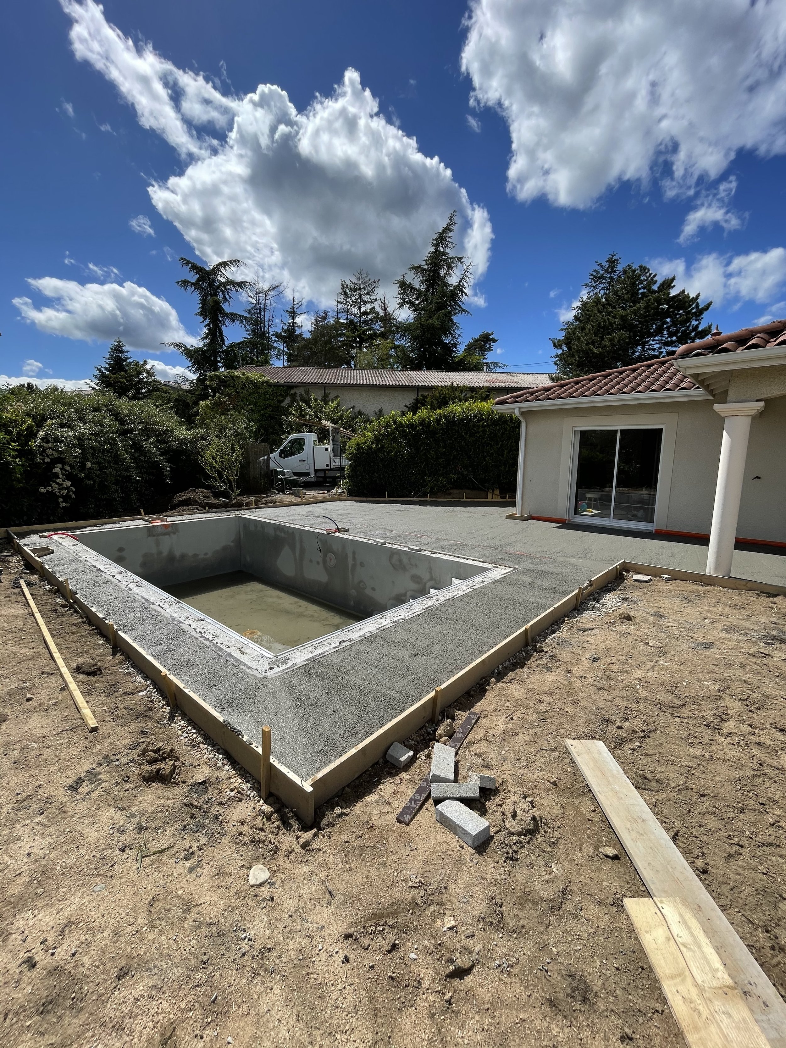 Une piscine en construction dans une cour arrière, entourée de terre et de quelques affiches de construction. Un bâtiment résidentiel avec un toit en tuiles est visible à l'arrière. Le ciel est bleu avec quelques nuages blancs.