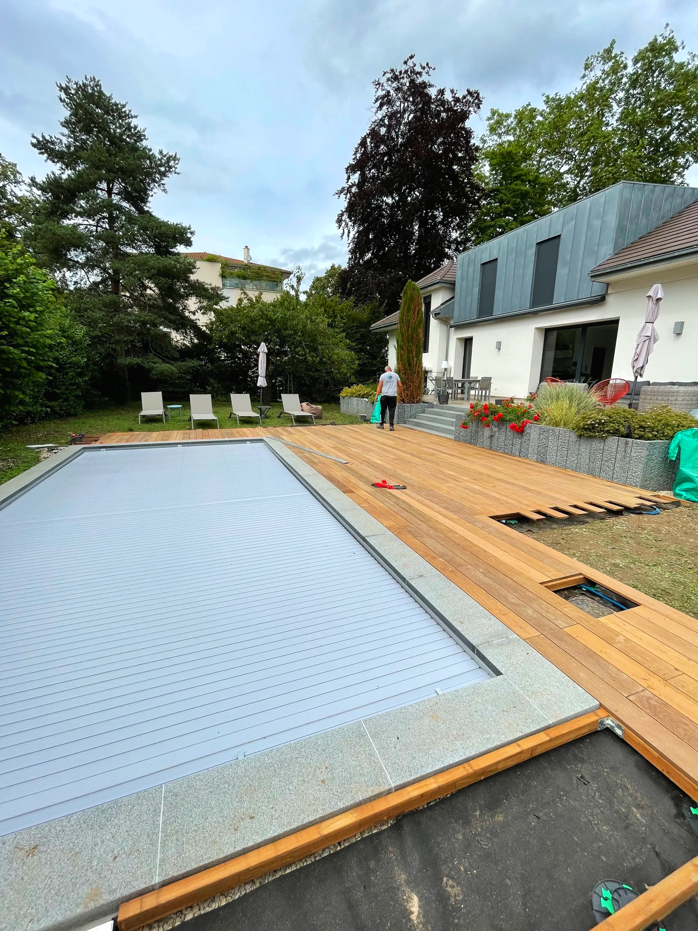 Travaux de rénovation de la terrasse d'une maison avec pose de lames de bois et couverture en toiture métallique, entourée d'arbres et de meubles de jardin.