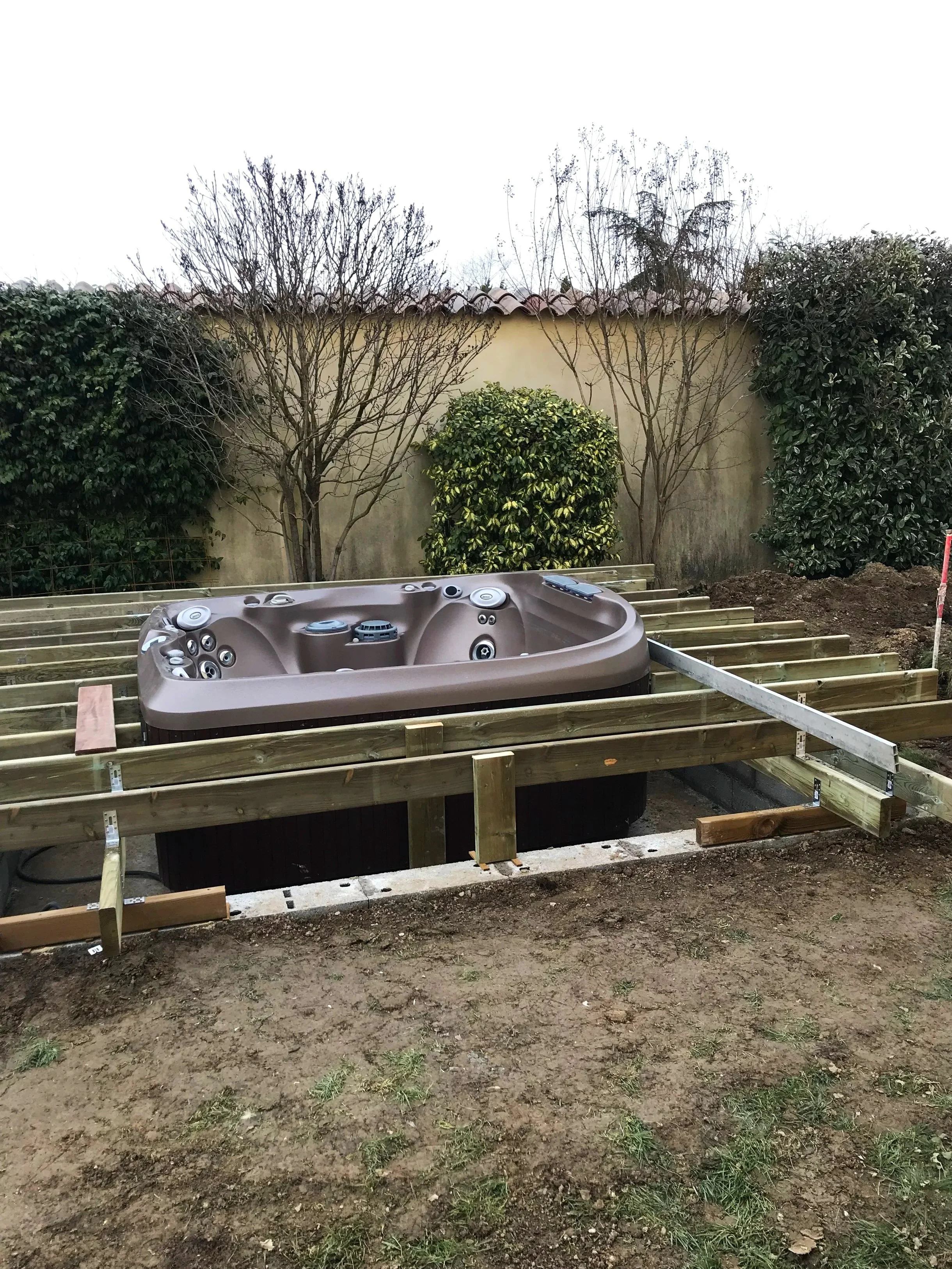 Une installation d'un spa dans un espace extérieur en cours de construction, entouré d'une structure en bois et de végétation, avec un mur en arrière-plan.