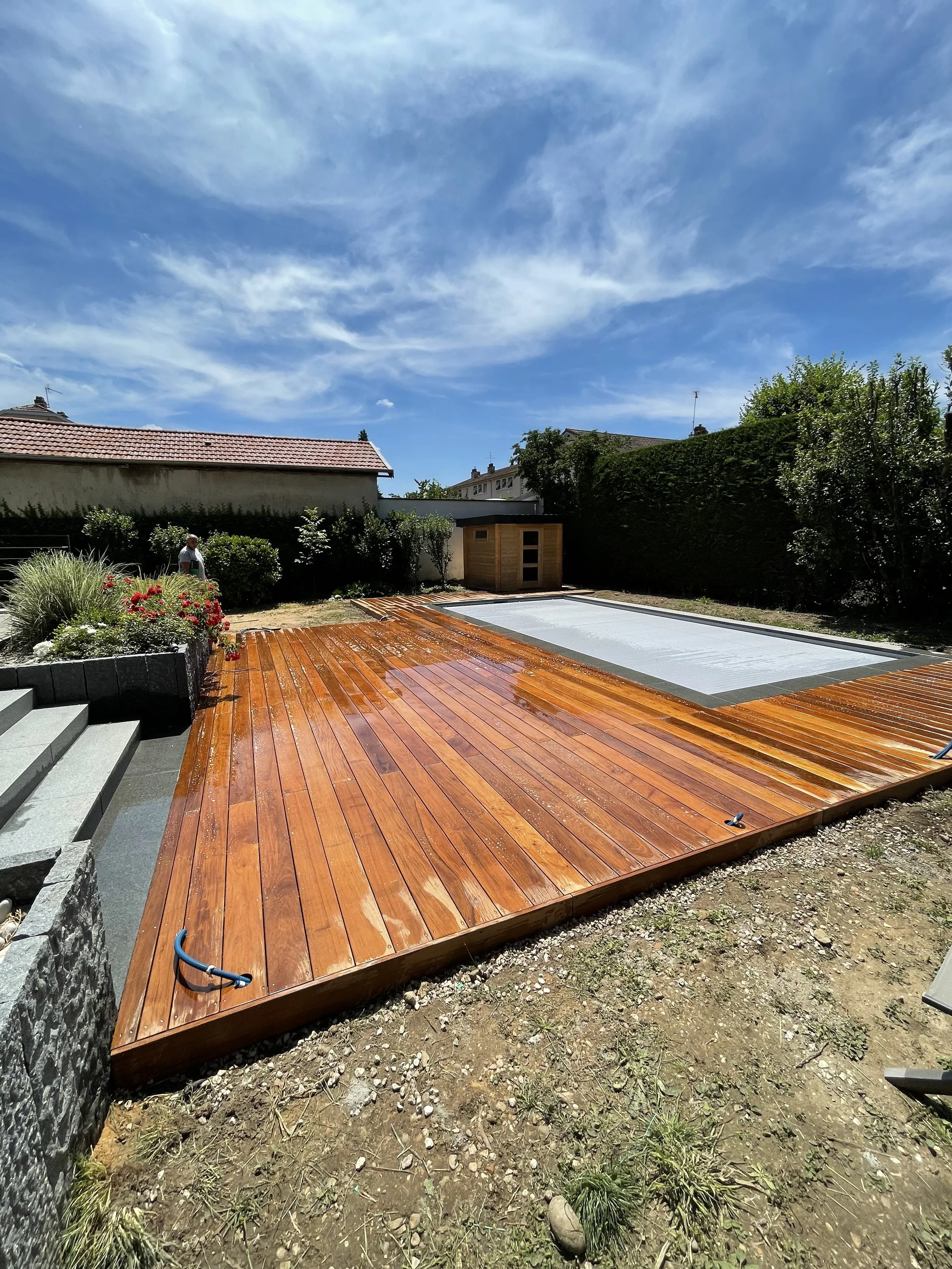 Un jardin avec une terrasse en bois fraîchement rénovée, un petit abri en bois, des plantes et un ciel bleu avec quelques nuages.