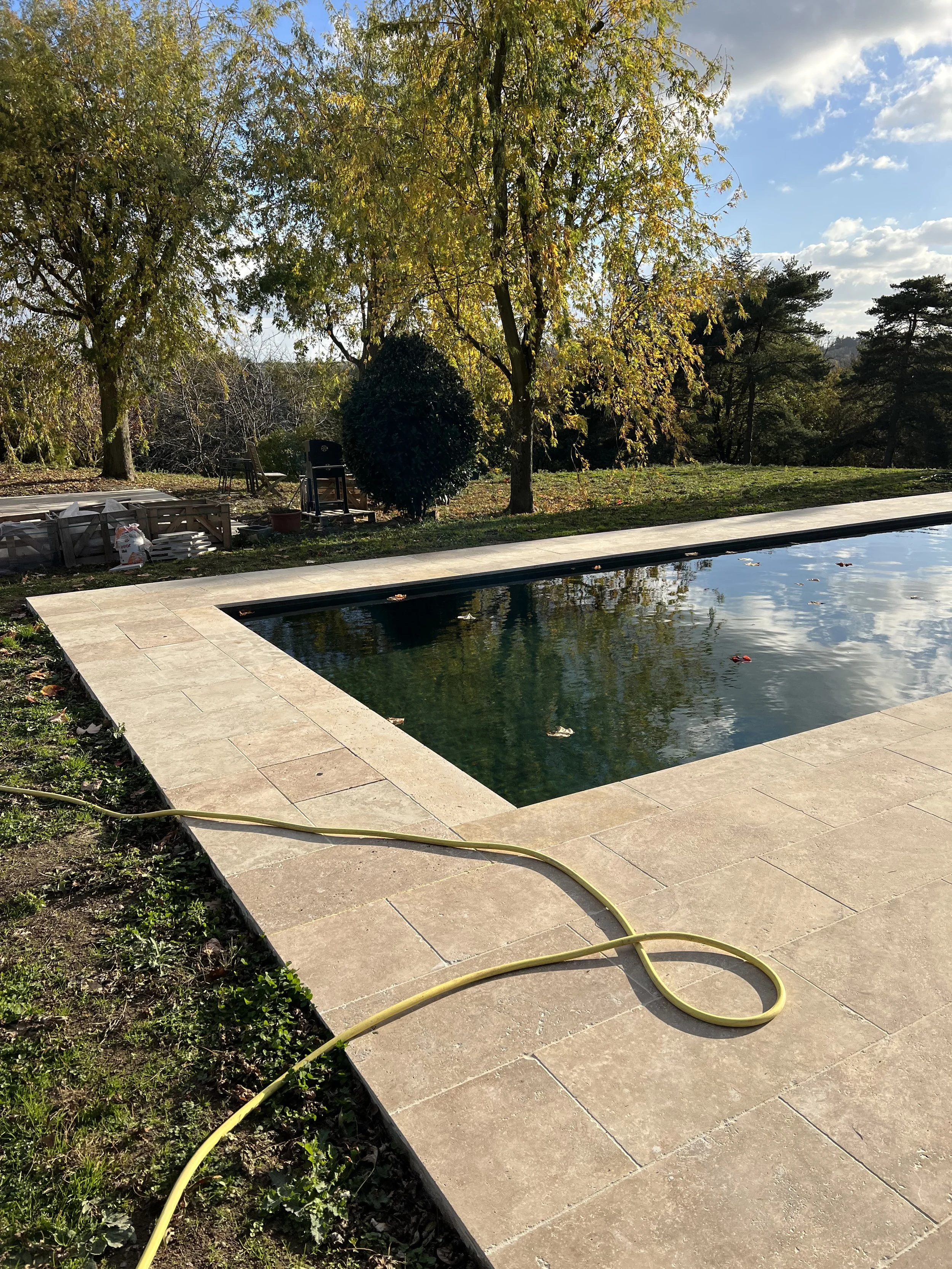Une piscine extérieure entourée de carreaux en pierre, avec des arbres verts et un ciel partiellement nuageux en arrière-plan, un tuyau d'arrosage jaune sur le sol.