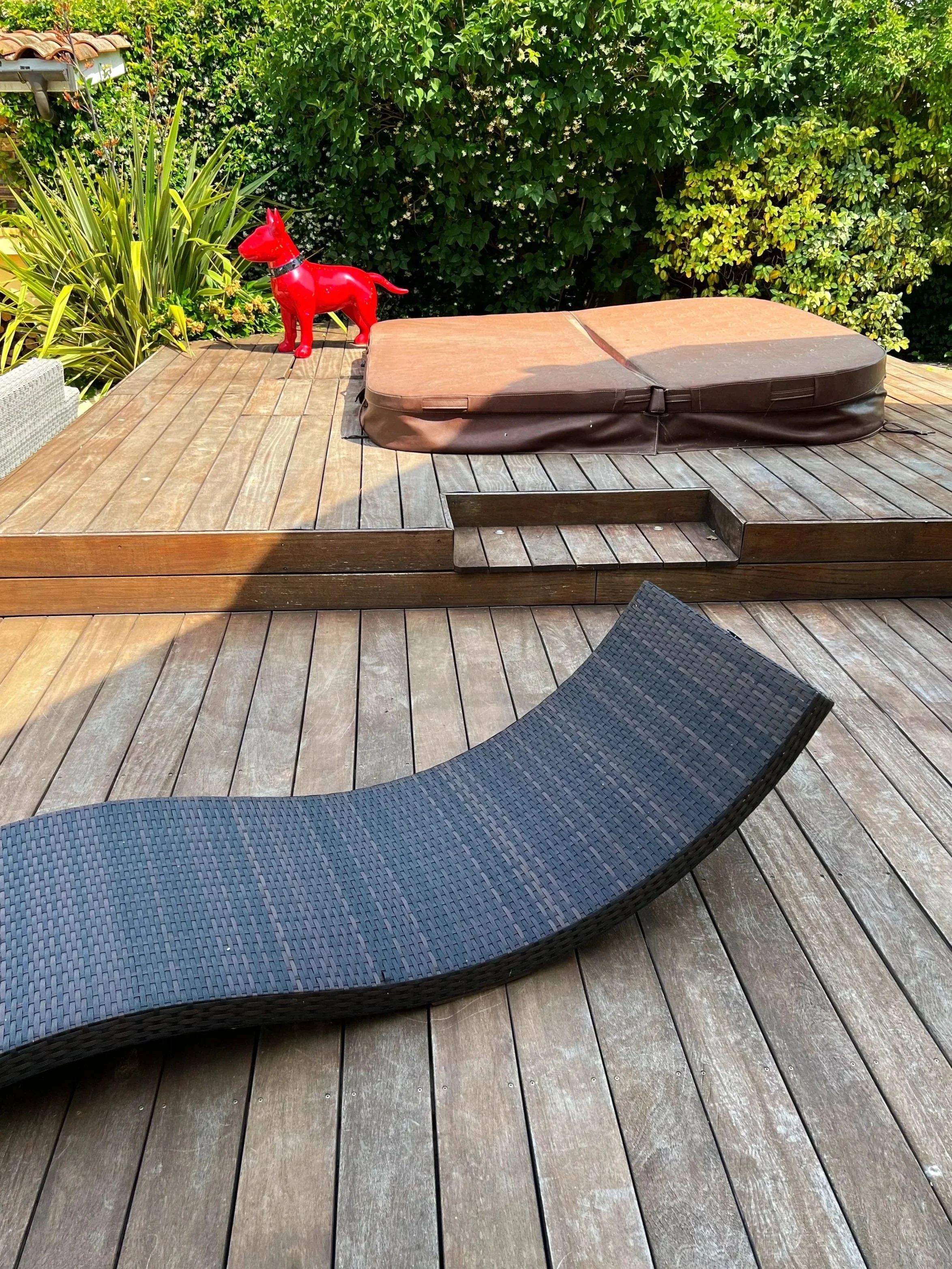 Chaise longue noire sur une terrasse en bois, avec un arbre vert en arrière-plan et une figurine de chien rouge près du coin de la terrace.