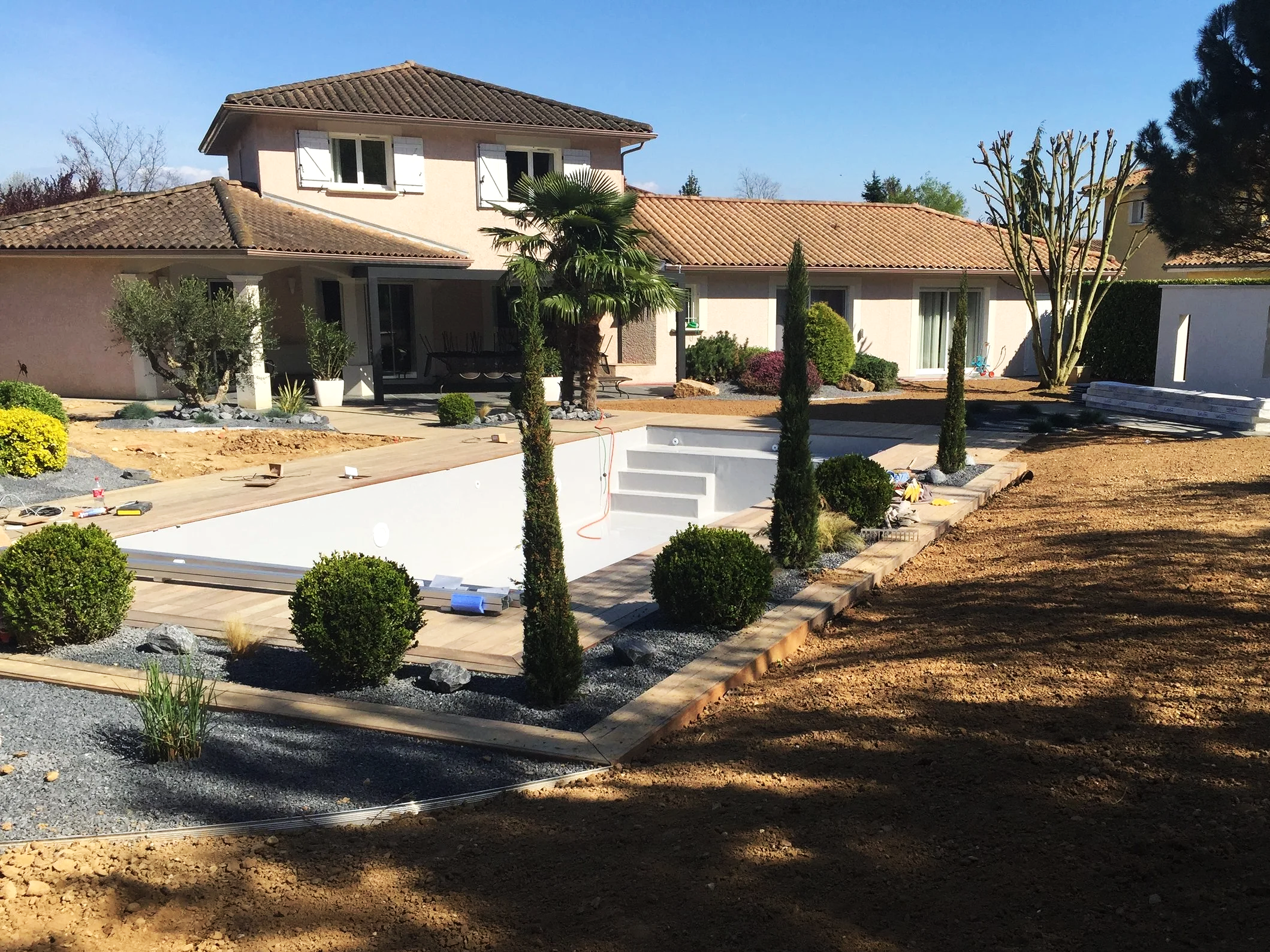 Une maison avec un jardin en cours d'aménagement, arbres, plantes et matériaux de construction visibles, sans humains.