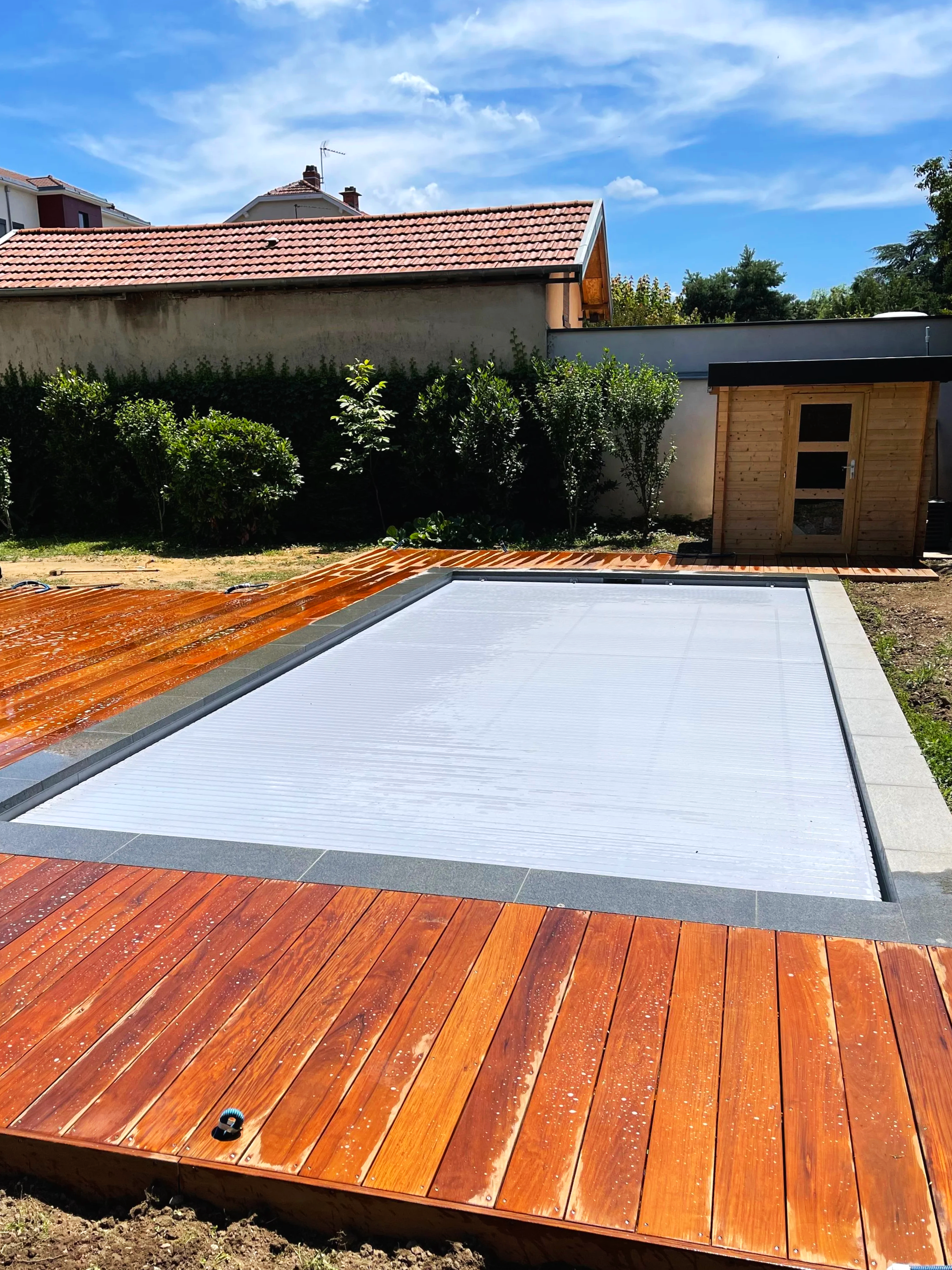 C'est une scène extérieure montrant une terrasse en bois avec un design en courbes, et une structure en bois, un mur blanc, des buissons verts, et des maisons derrière avec un ciel bleu et nuages blancs.