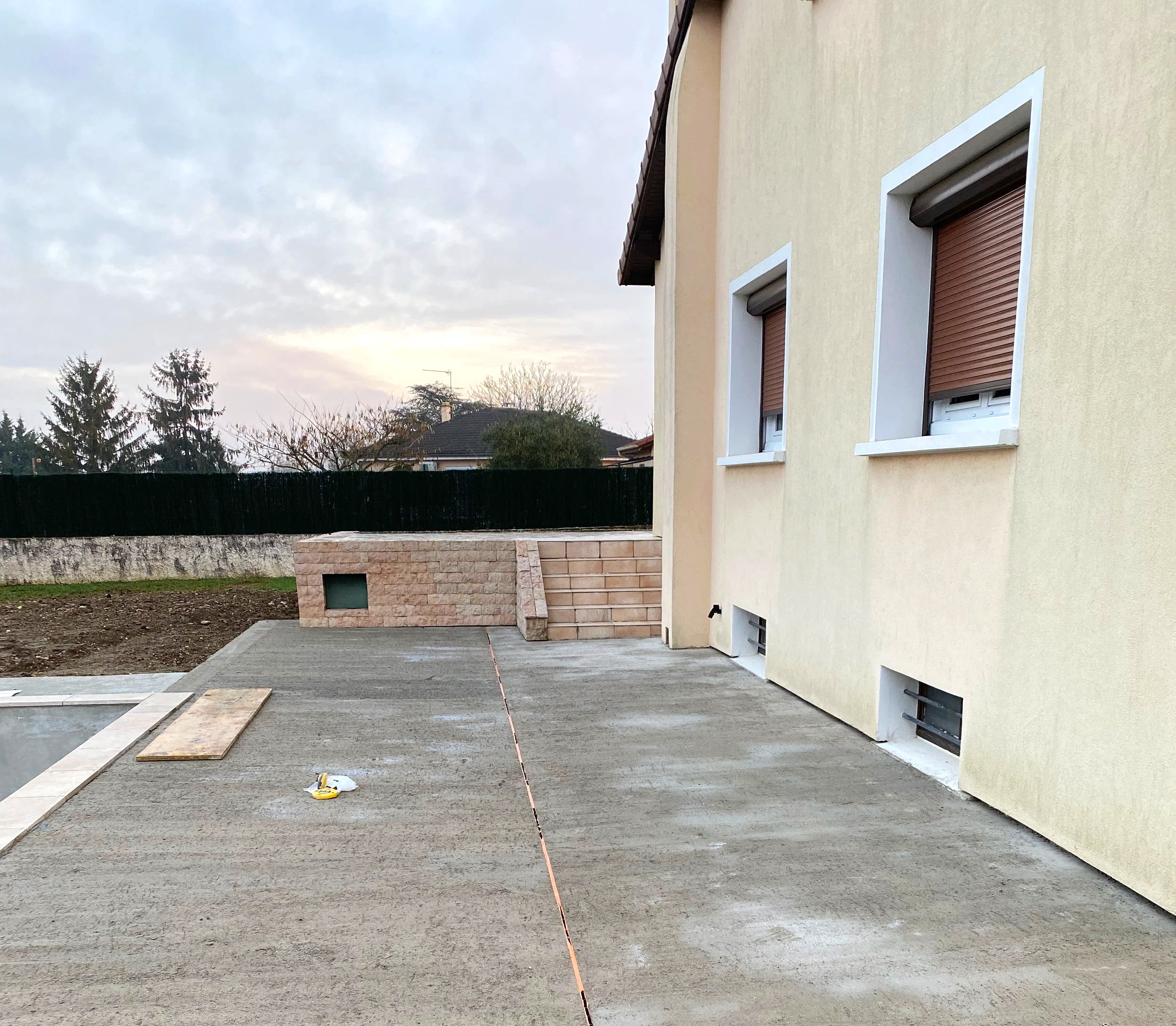 Une maison avec des fenêtres à volets roulants, en cours de construction ou de rénovation, avec un espace extérieur en gravier et des plans ou des matériaux de construction sur le sol.
