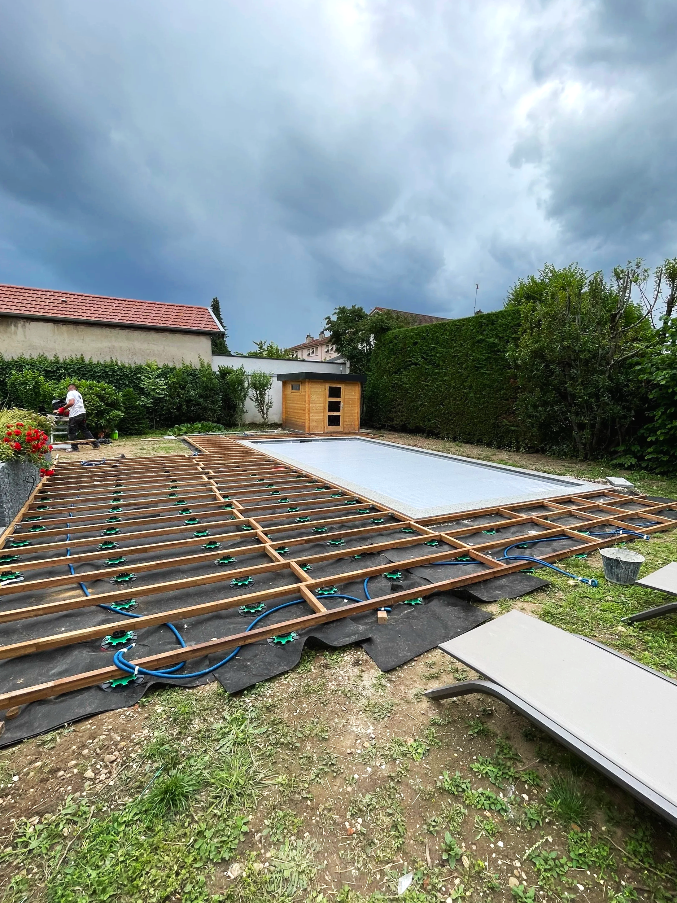 Travaux de construction d'une piscine avec planches en bois, tuyaux, et un abri en bois dans un jardin. Ciel nuageux en arrière-plan.