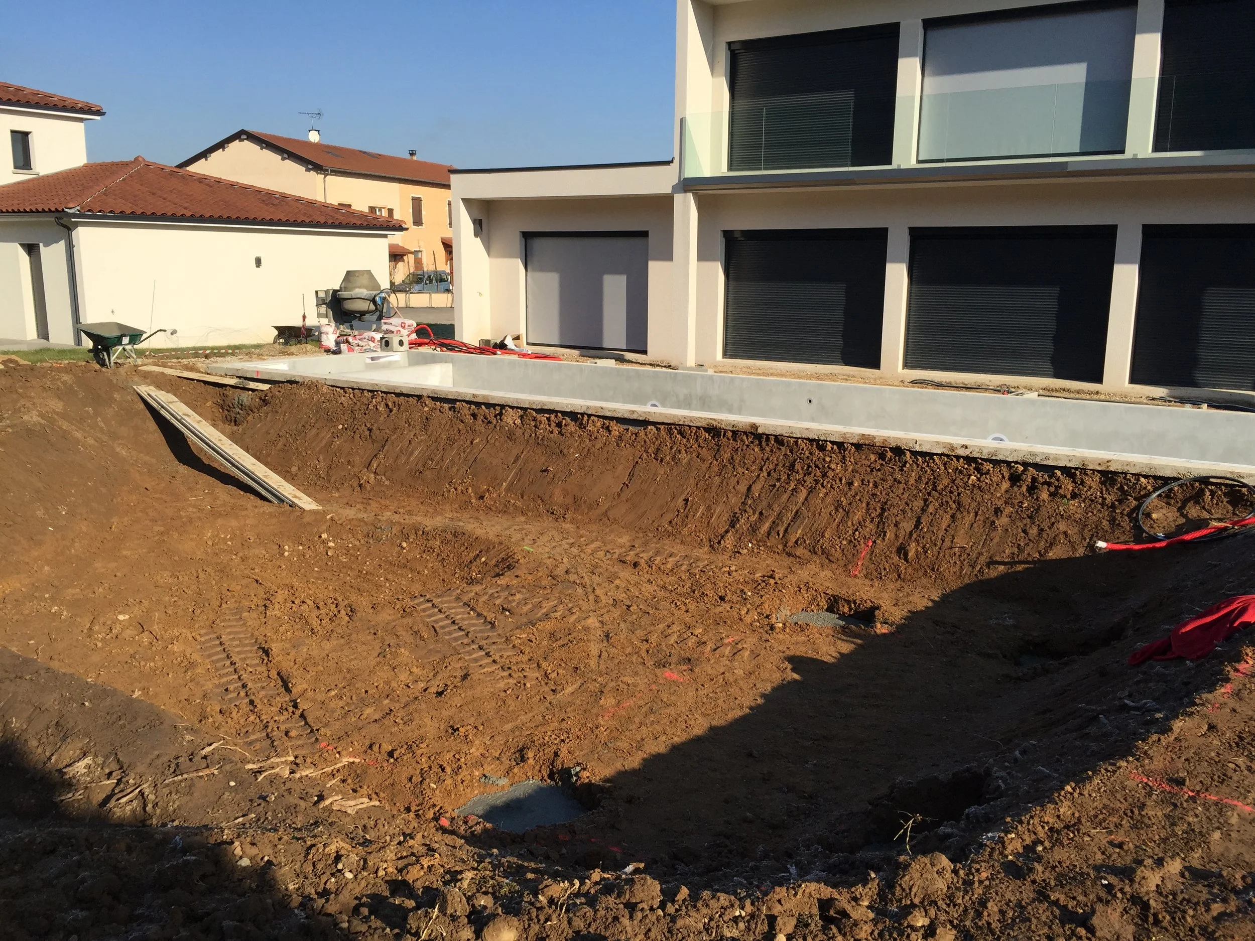 Un bâtiment moderne en construction avec une excavation en cours devant. La façade du bâtiment est blanche avec des fenêtres à volets roulants noirs. Il y a des outils et des matériaux de construction posés sur le site.