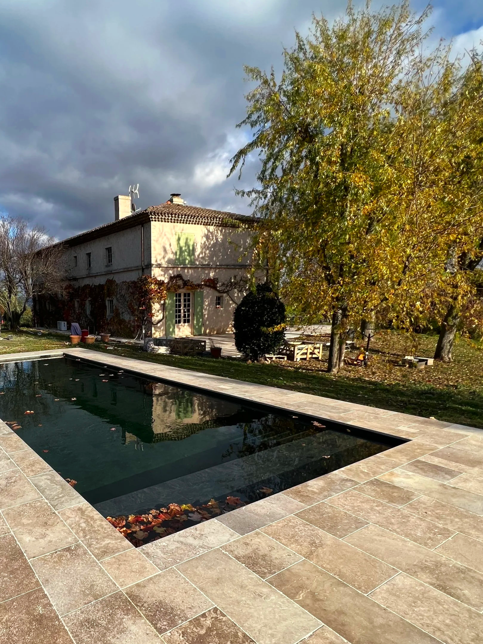 Une piscine rectangulaire en pierre dans un jardin avec une maison en arrière-plan, des arbres avec des feuilles automnales et un ciel nuageux.
