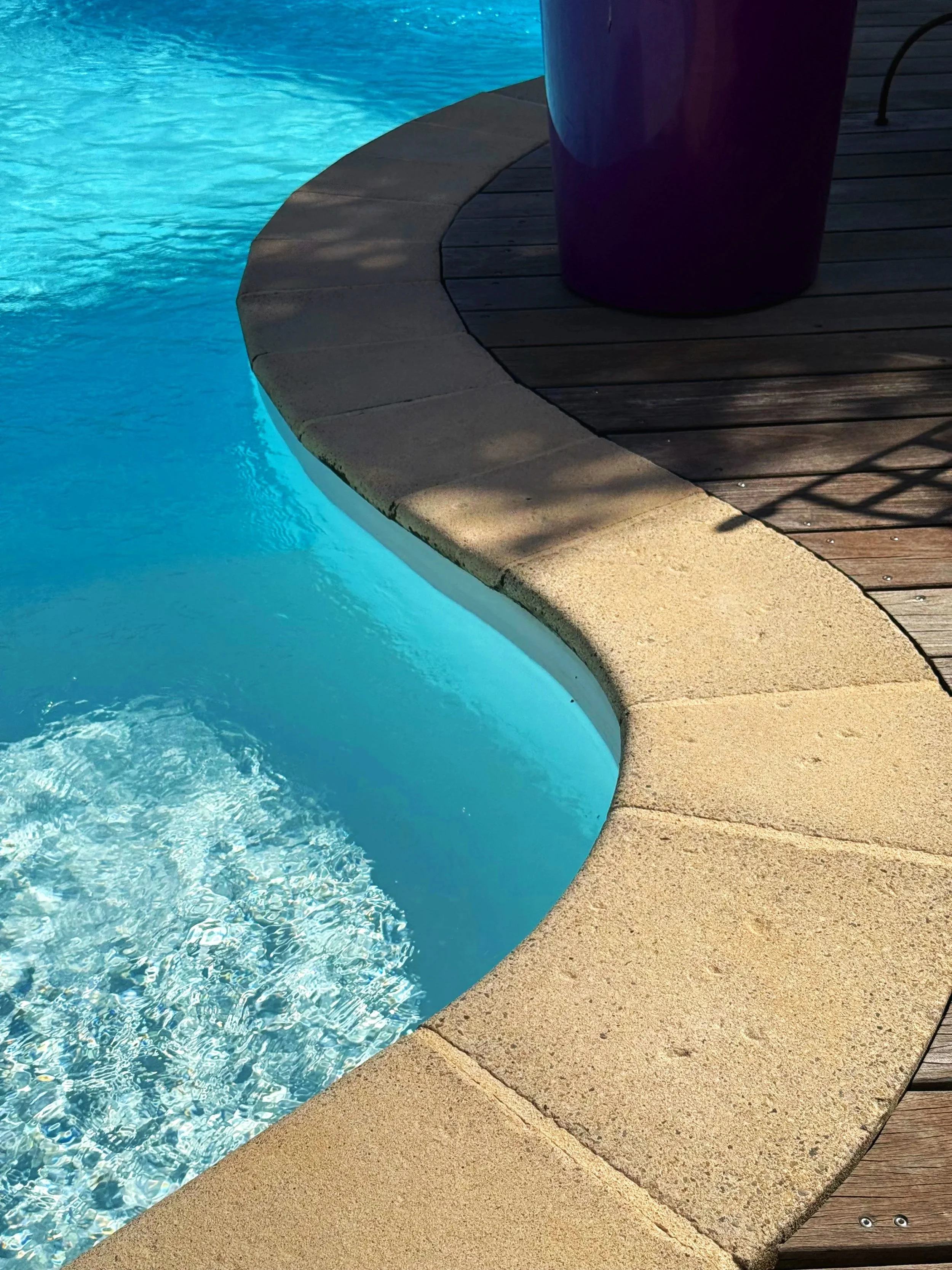 Bord de piscine avec eau bleue claire, margelles en pierre, surface en bois à proximité, partie d'un grand pot violet.