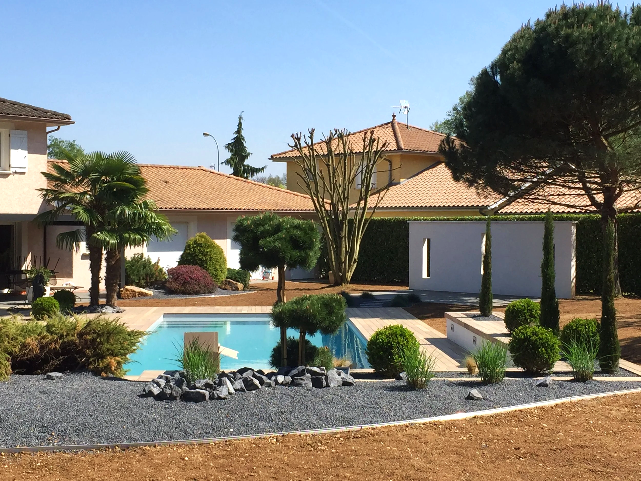 Une maison avec un toit en tuiles, un jardin bien entretenu, une piscine, et plusieurs arbres, dont un arbre grand et feuillu à droite et un palmier à gauche. Le ciel est clair et ensoleillé.