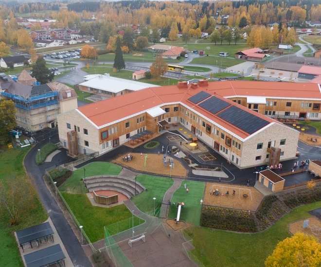 Låg flygplansvinkelbild av en skolbyggnad med lekplatser och omgivande grönområden under höst. Vinterbilder av skolans tak med solceller.