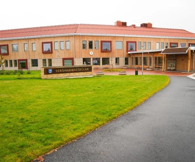 En skolbyggnad av trä med ett grönt gräsmattem och en asfalterad väg framför. En skylt med texten 'RENSHAMMAR SKOLAN'.